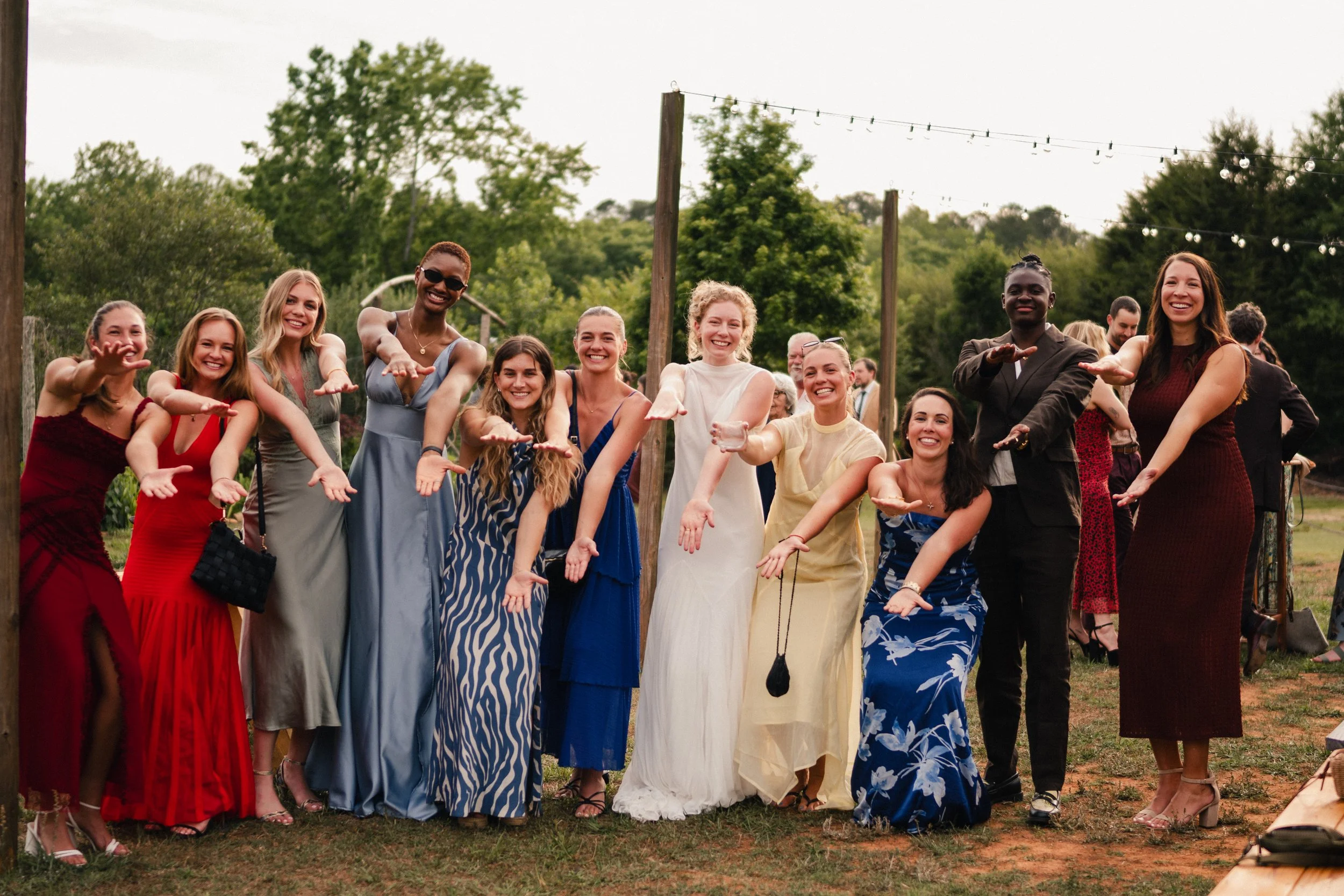A group of women and men smiling and extending their arms forward at an outdoor event with string lights and trees in the background.