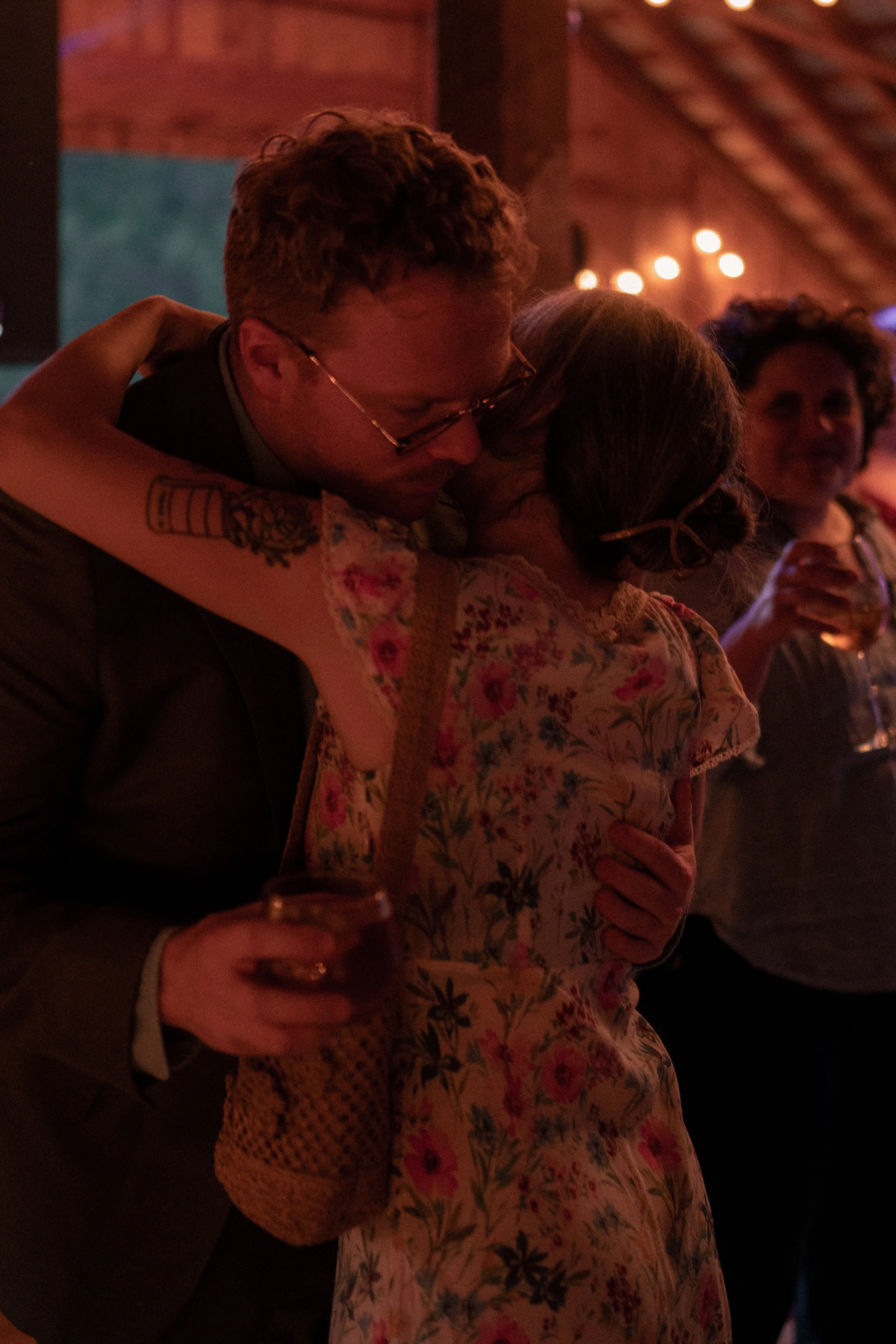 A couple hugging tightly in a dimly lit setting, with a person holding a glass nearby. The atmosphere appears warm and intimate, possibly at a social gathering or party.