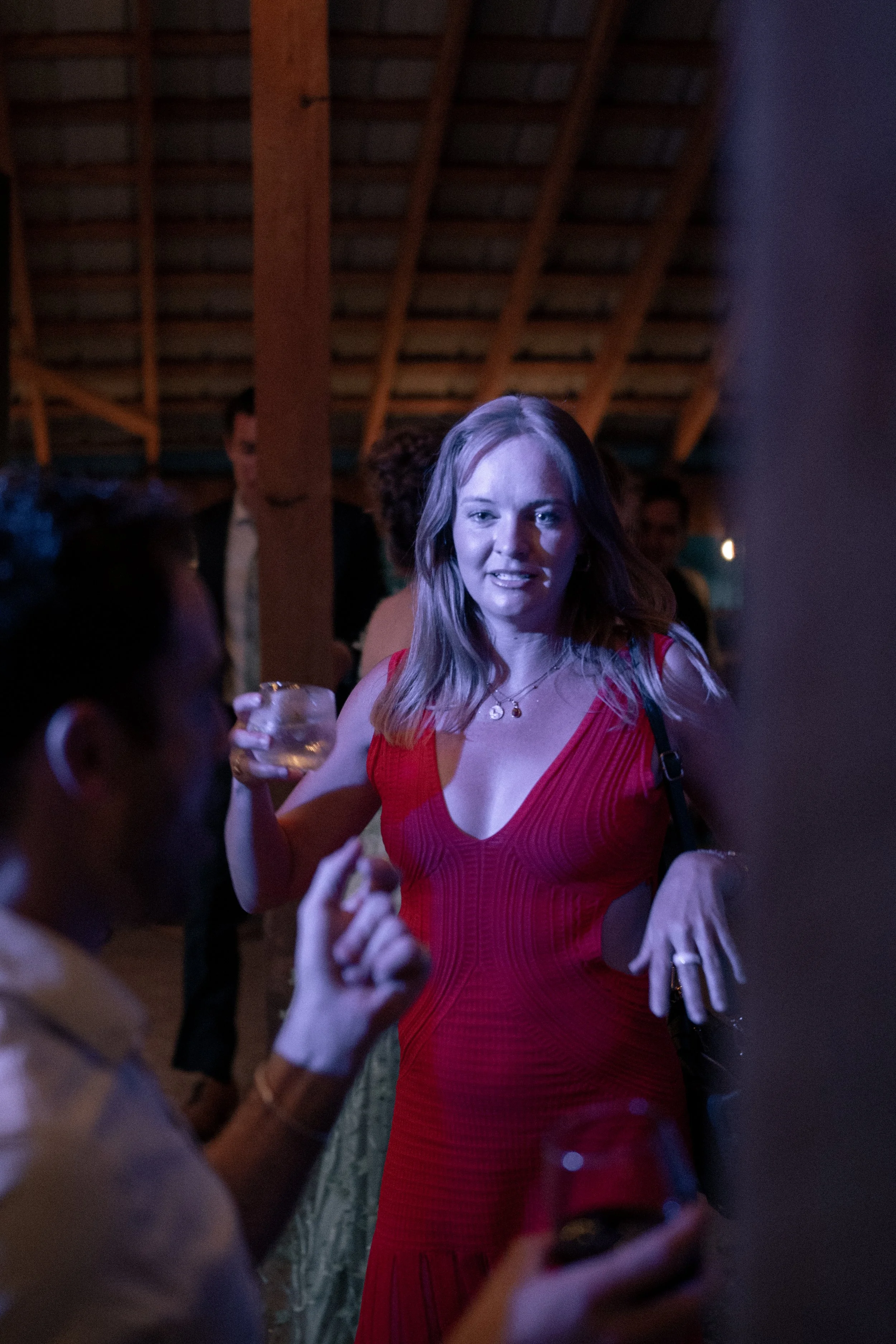 A woman in a red dress at a party, holding a drink, talking to a man. The setting appears to be a dimly lit indoor venue with a wooden ceiling and other people in the background.
