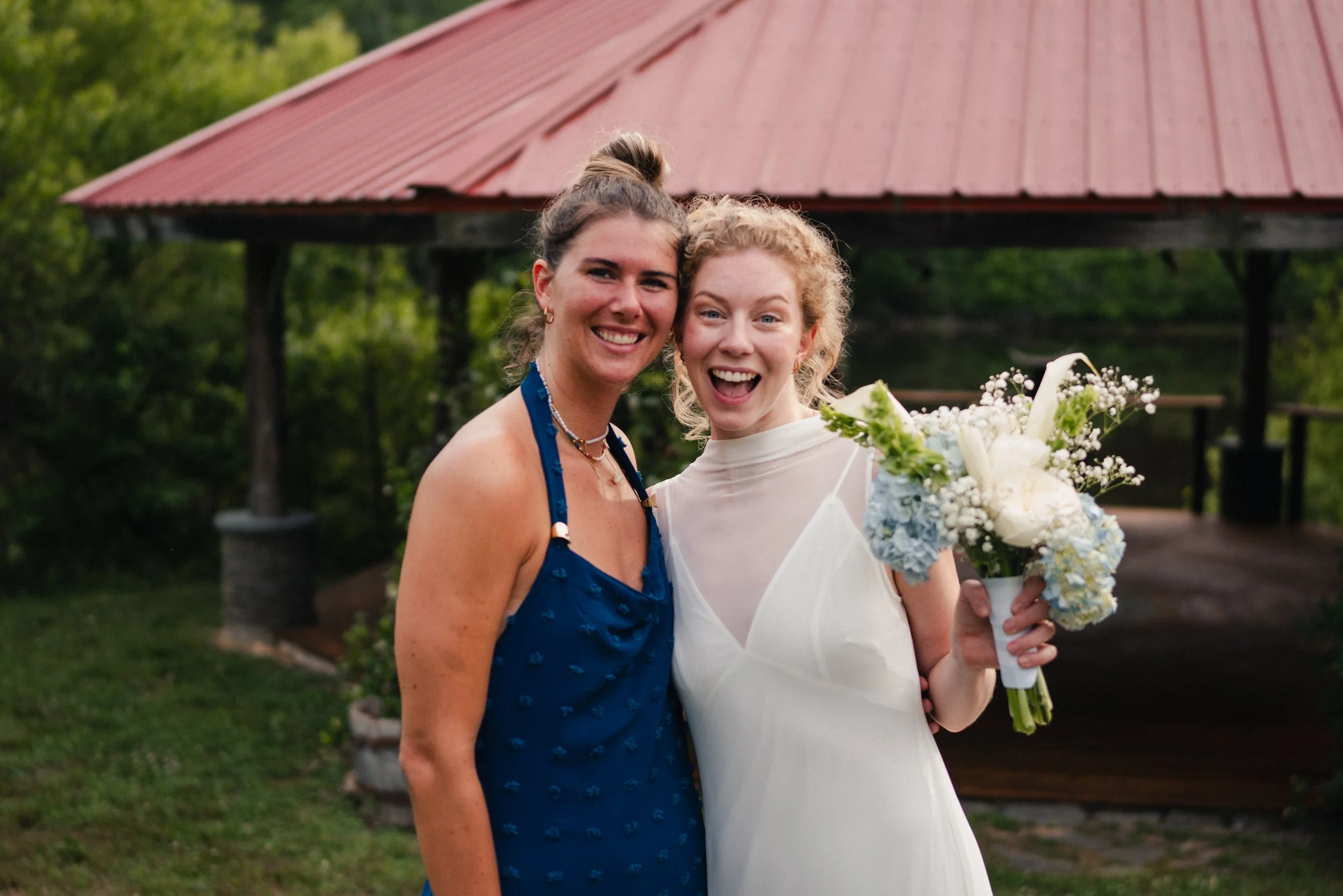 Two women, one with curly red hair in a white dress, holding a bouquet of white and blue flowers, smiling outdoors with a red-roofed pavilion and green trees in the background.