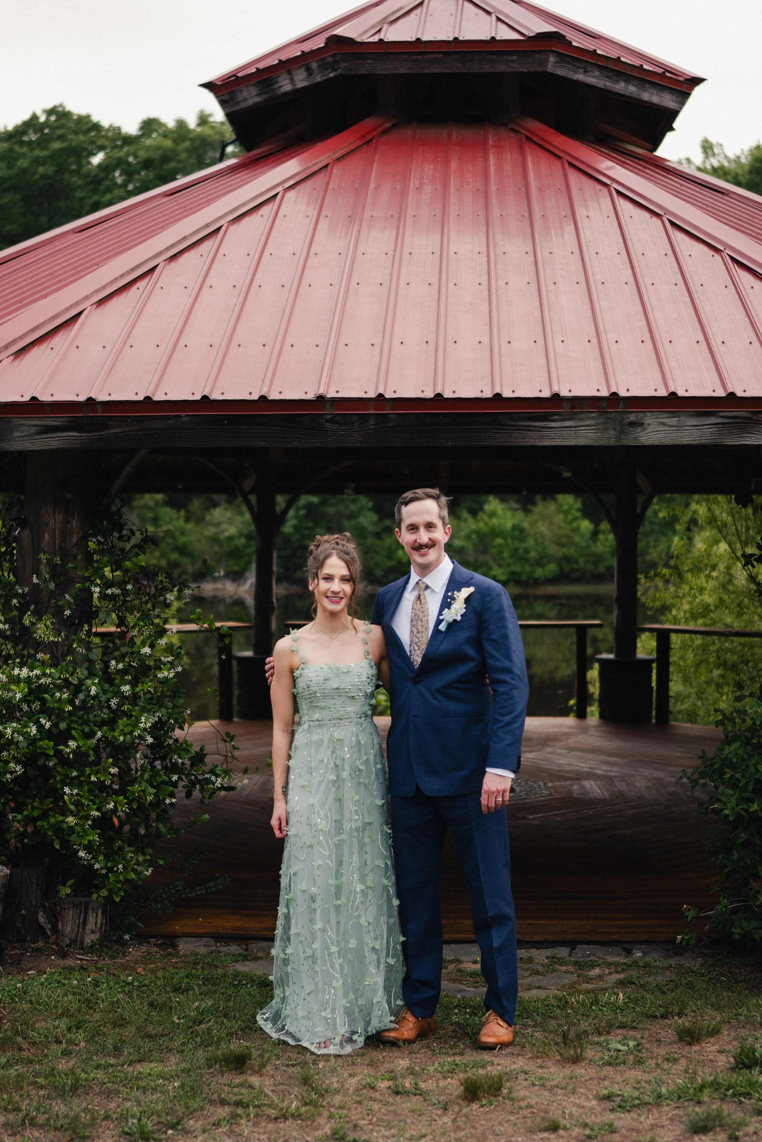 A couple dressed in formal attire standing under a pavilion with a red metal roof, surrounded by greenery.