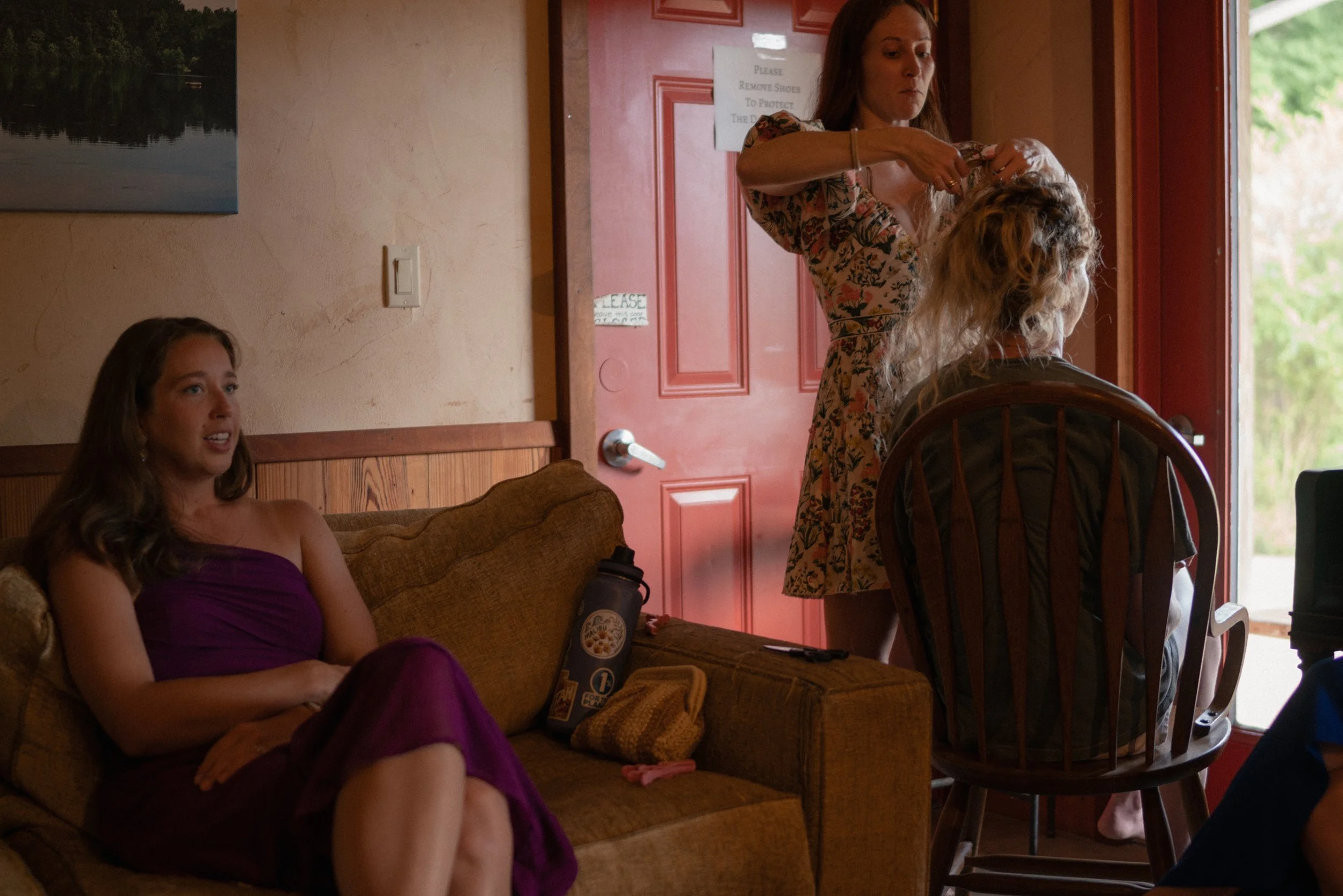 A woman with long brown hair, wearing a floral dress, is standing behind a seated woman with curly blonde hair, who is facing away. The standing woman appears to be styling or adjusting the seated woman's hair. Another woman in a purple outfit is sit
