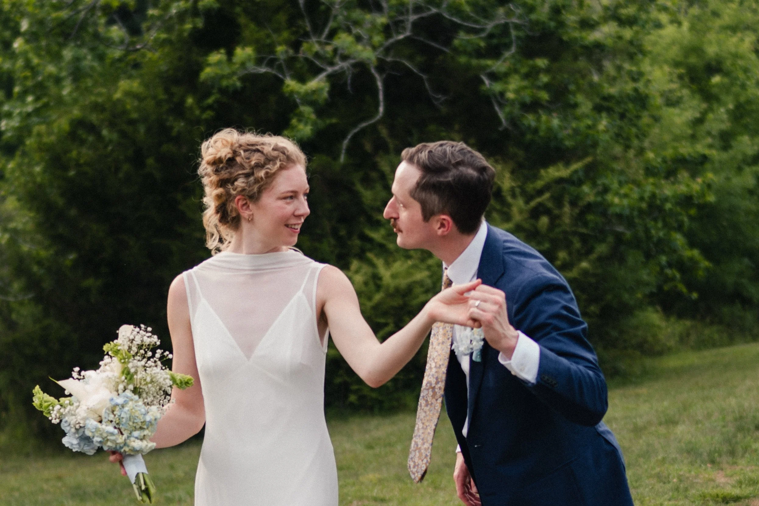 A bride and groom dancing outdoors with trees in the background. The bride is holding a bouquet of flowers, and they are smiling at each other.