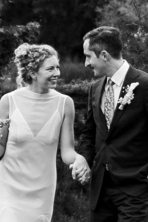 A smiling woman and a man in a suit holding hands outdoors, likely at a wedding or special event.