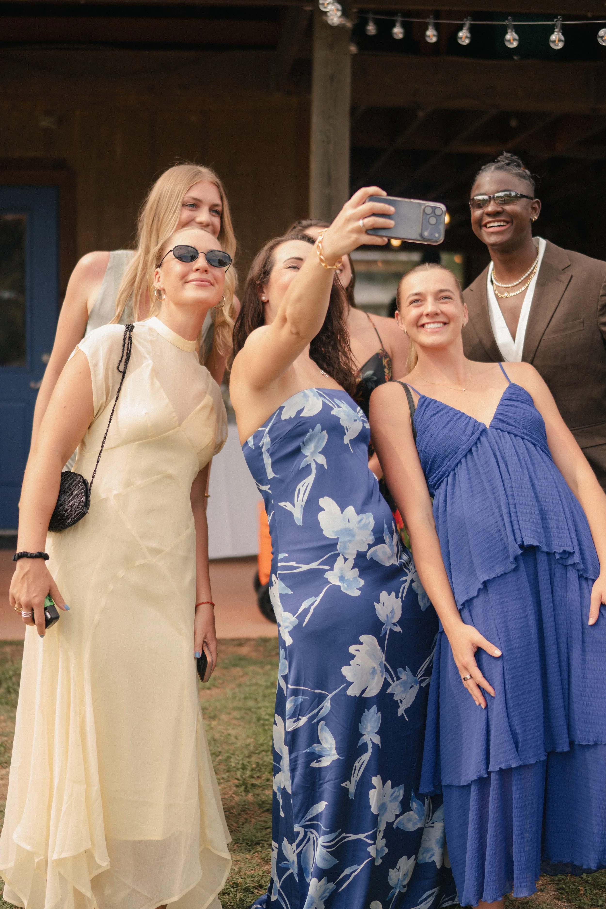 A group of six women taking a selfie together at an outdoor event, dressed in colorful, elegant dresses, with string lights overhead and a wooden structure in the background.