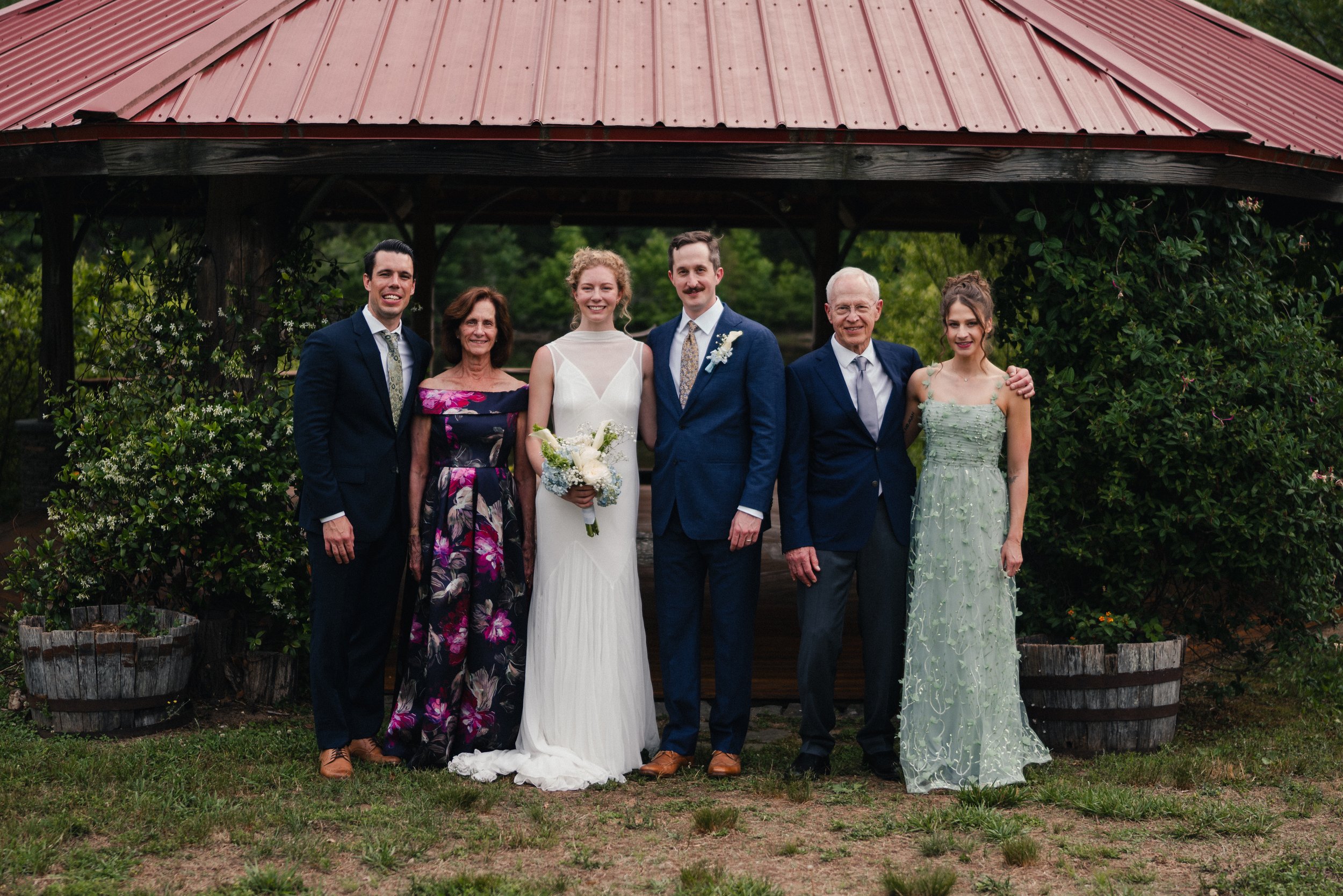 Group of six people at a wedding, standing in front of a wooden gazebo with a red metal roof, surrounded by greenery.