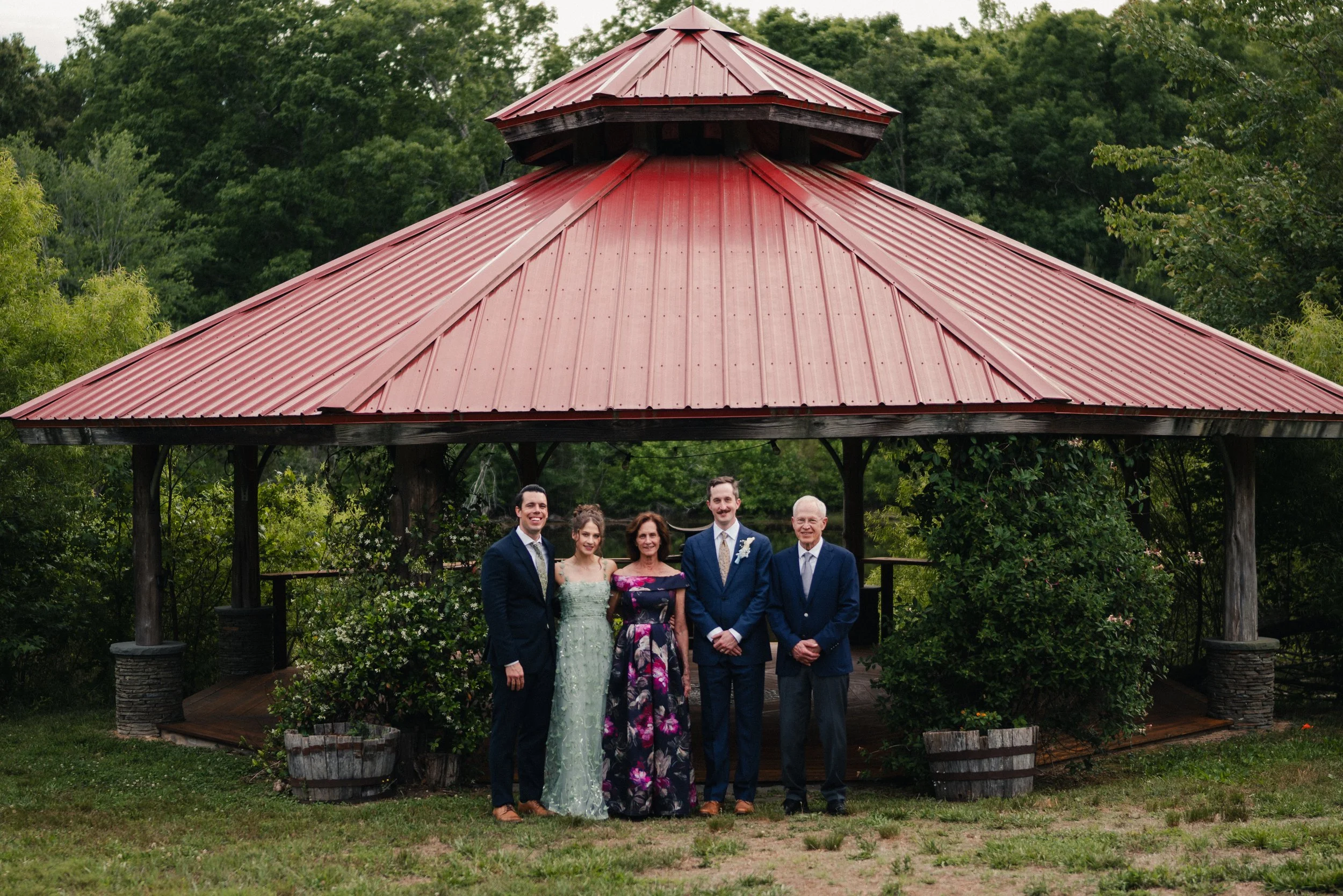 Group of six people, including men in suits and women in formal dresses, standing together outdoors under a gazebo with a red metal roof, surrounded by trees and greenery.