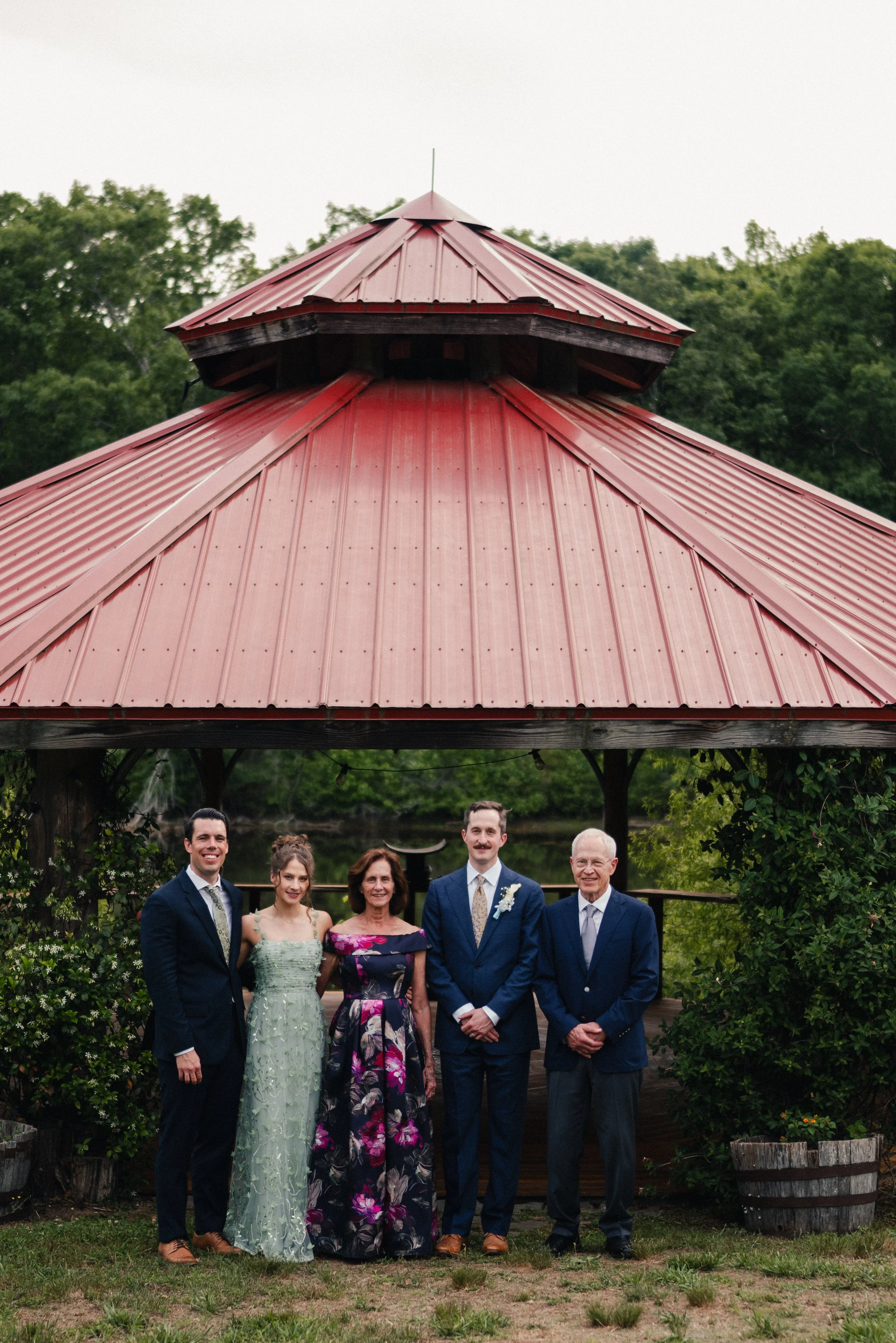 A family gathering outdoors under a red gazebo with green trees in the background. Five adults in formal attire are standing side by side, smiling at the camera. The women are wearing dresses, and the men are in suits.