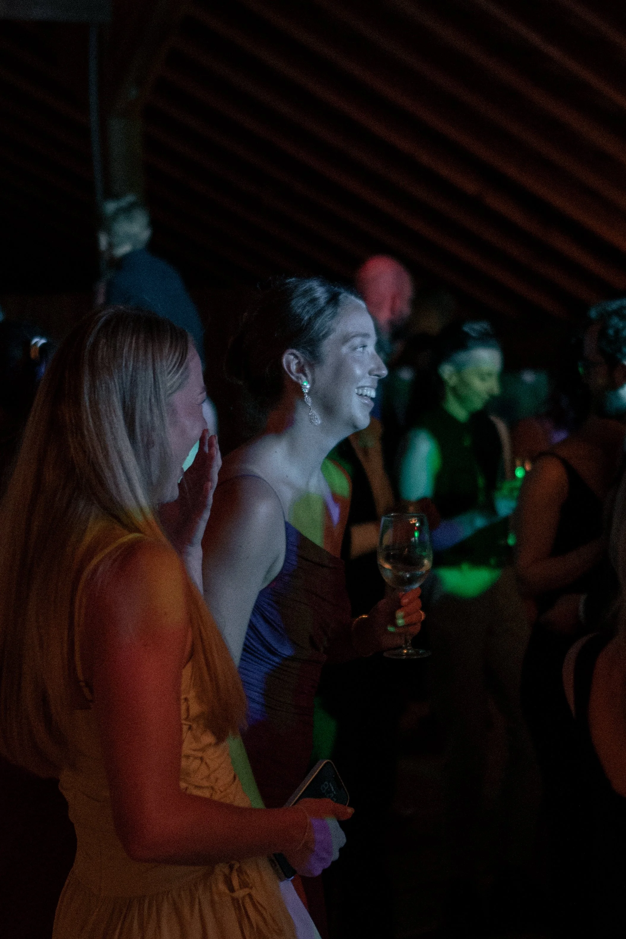 People dressed in colorful evening attire, smiling and engaging at a social event in a dimly lit venue with a wooden ceiling.
