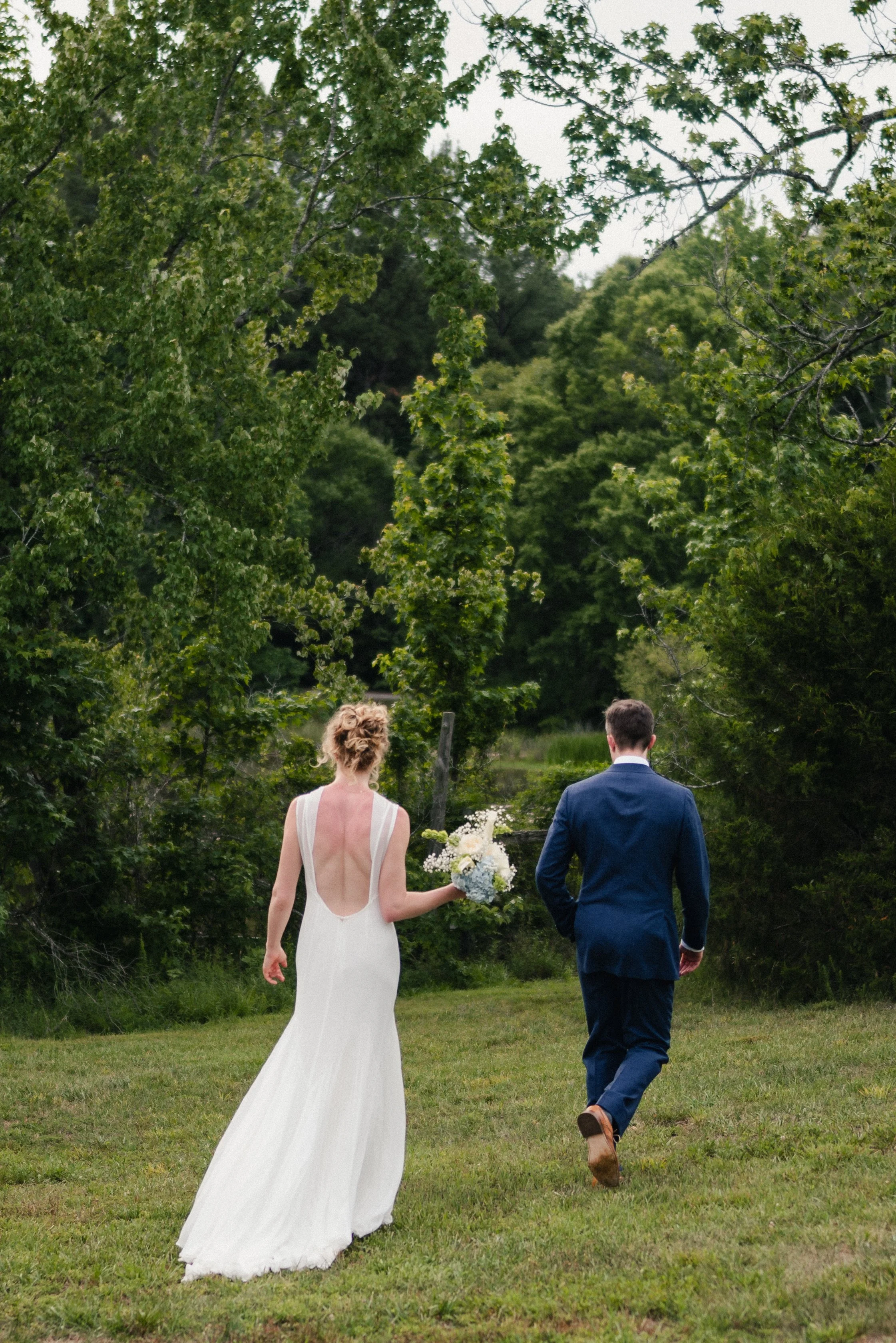 A bride and groom walk away outdoors in a grassy area surrounded by green trees. The bride wears a white gown with a low back, and the groom wears a dark suit. The bride holds a bouquet of flowers.