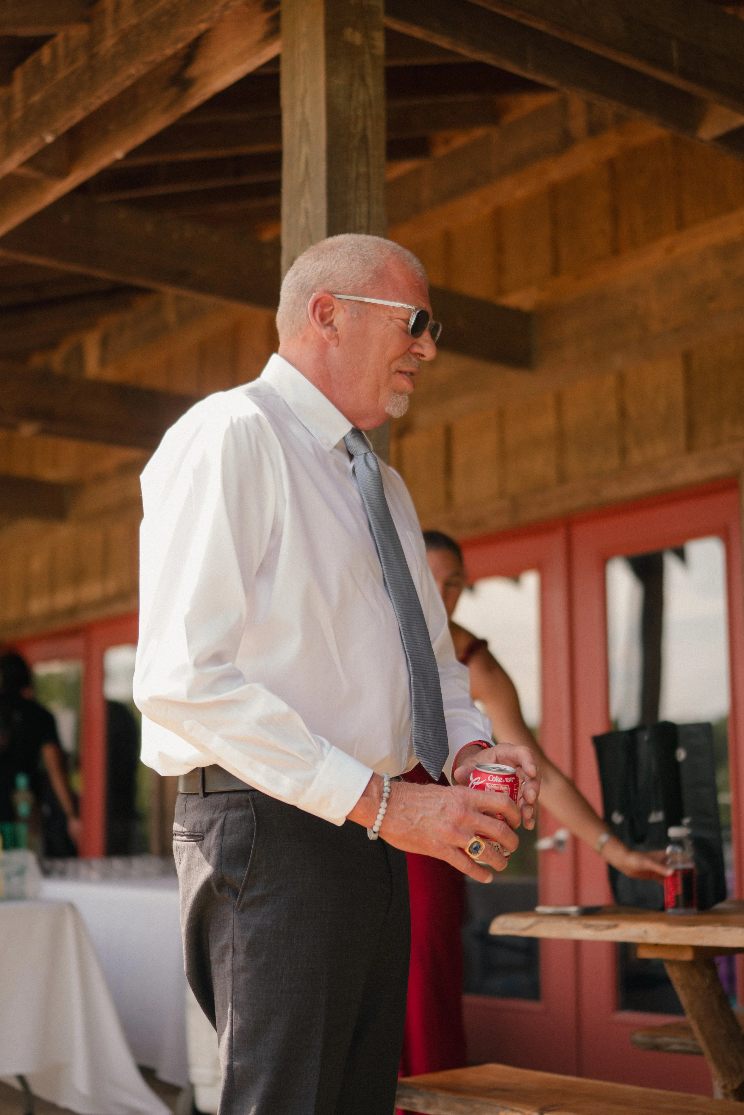 A man with a shaved head, sunglasses, and a gray tie, wearing a white dress shirt and dark pants, holding a Coca-Cola can at an outdoor event under a wooden pavilion.