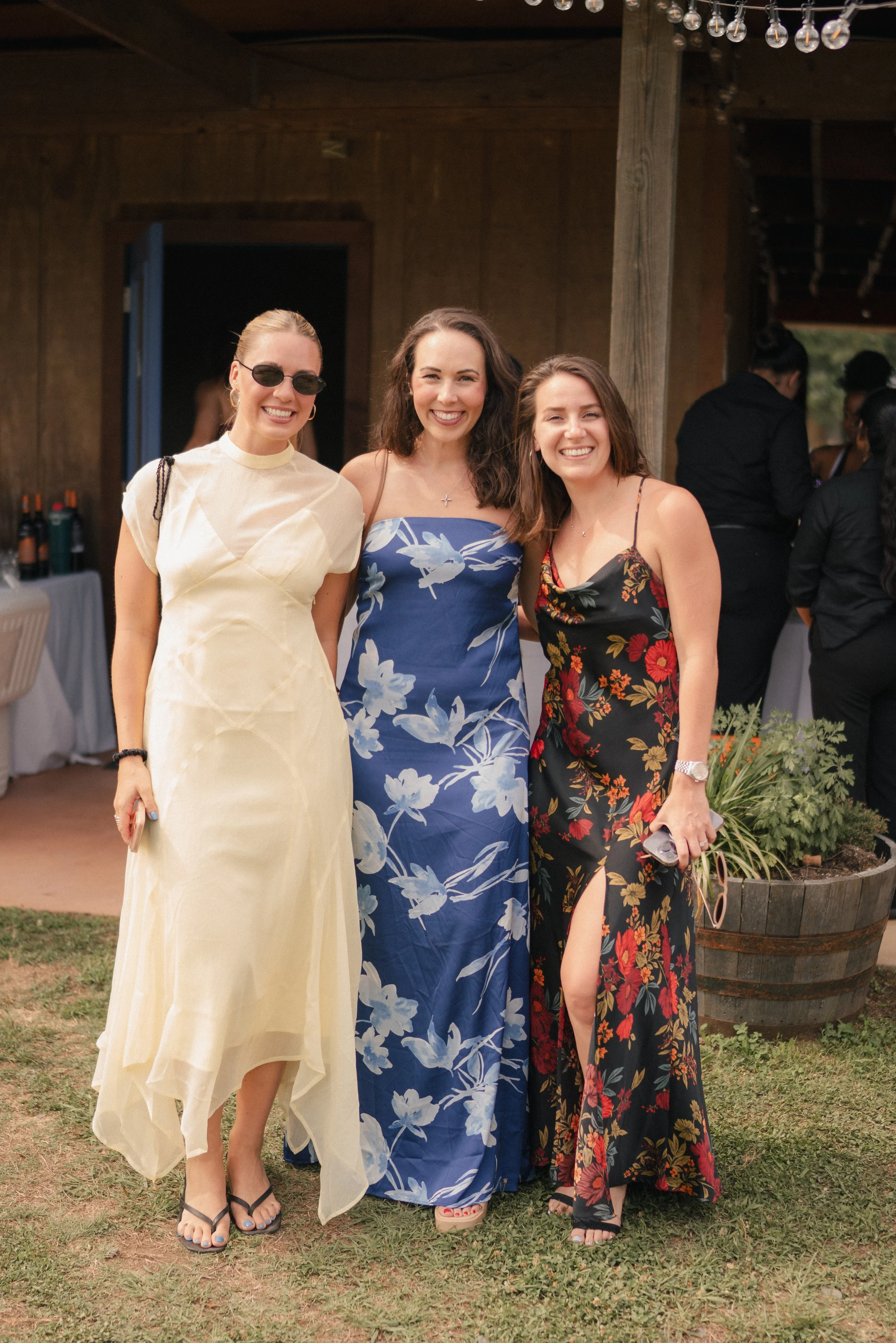Three women smiling and posing outdoors, dressed in colorful summer dresses, with a wooden building and other people in the background.