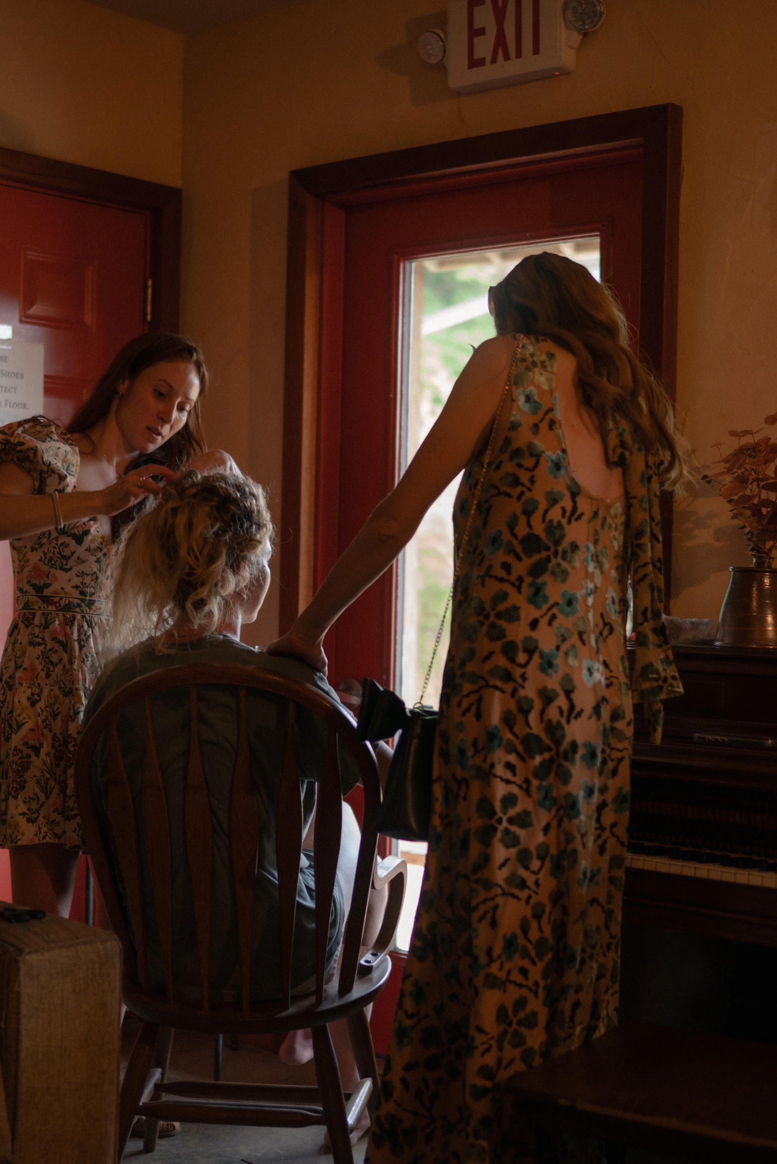 Two women with long hair are in a room near a red door, one is sitting in a wooden chair and the other is standing next to her. They are both wearing floral dresses, and seem to be engaged in a conversation or activity involving the seated woman. The