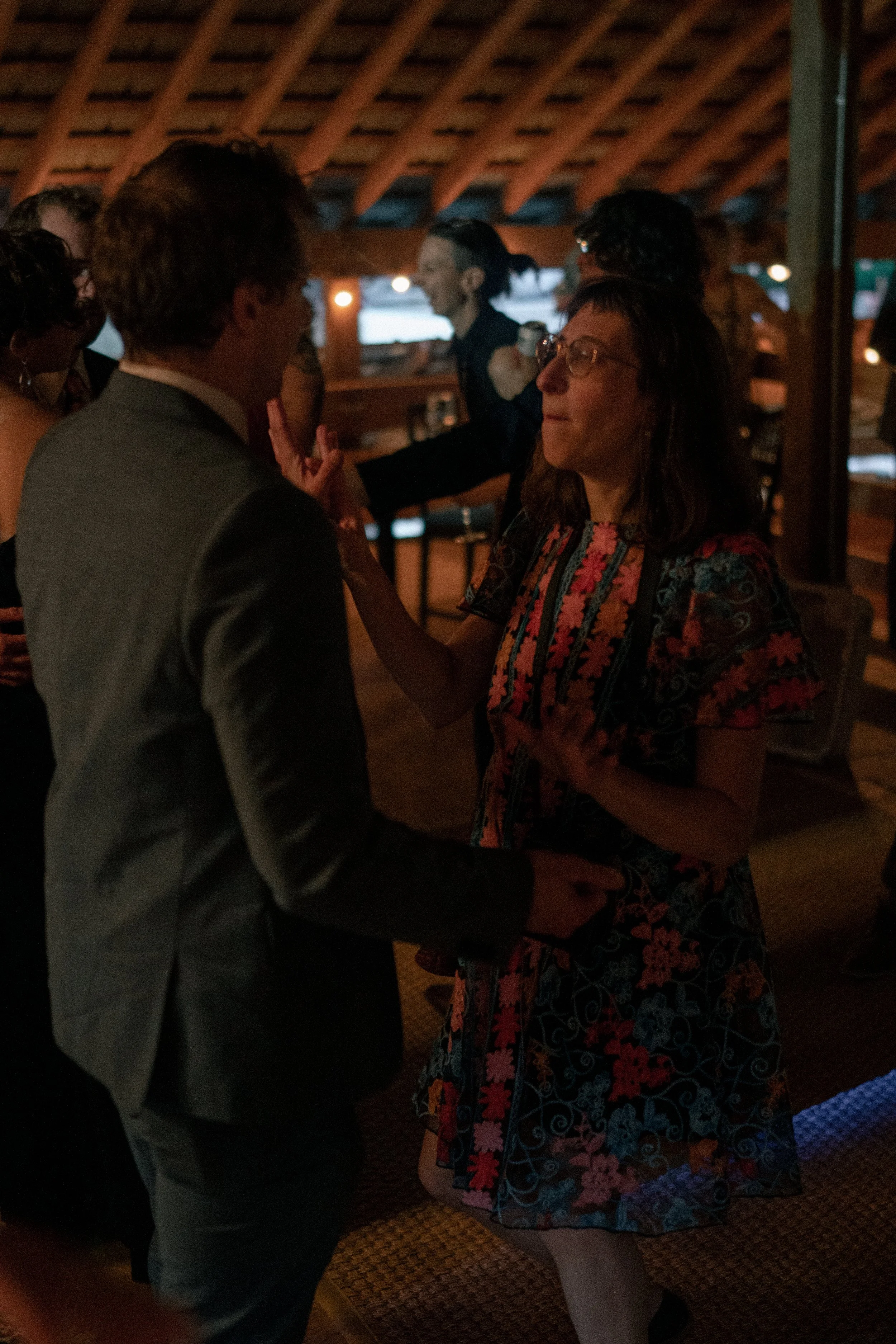 A man in a suit and a woman in a colorful dress are dancing closely together at an indoor social gathering with warm lighting.