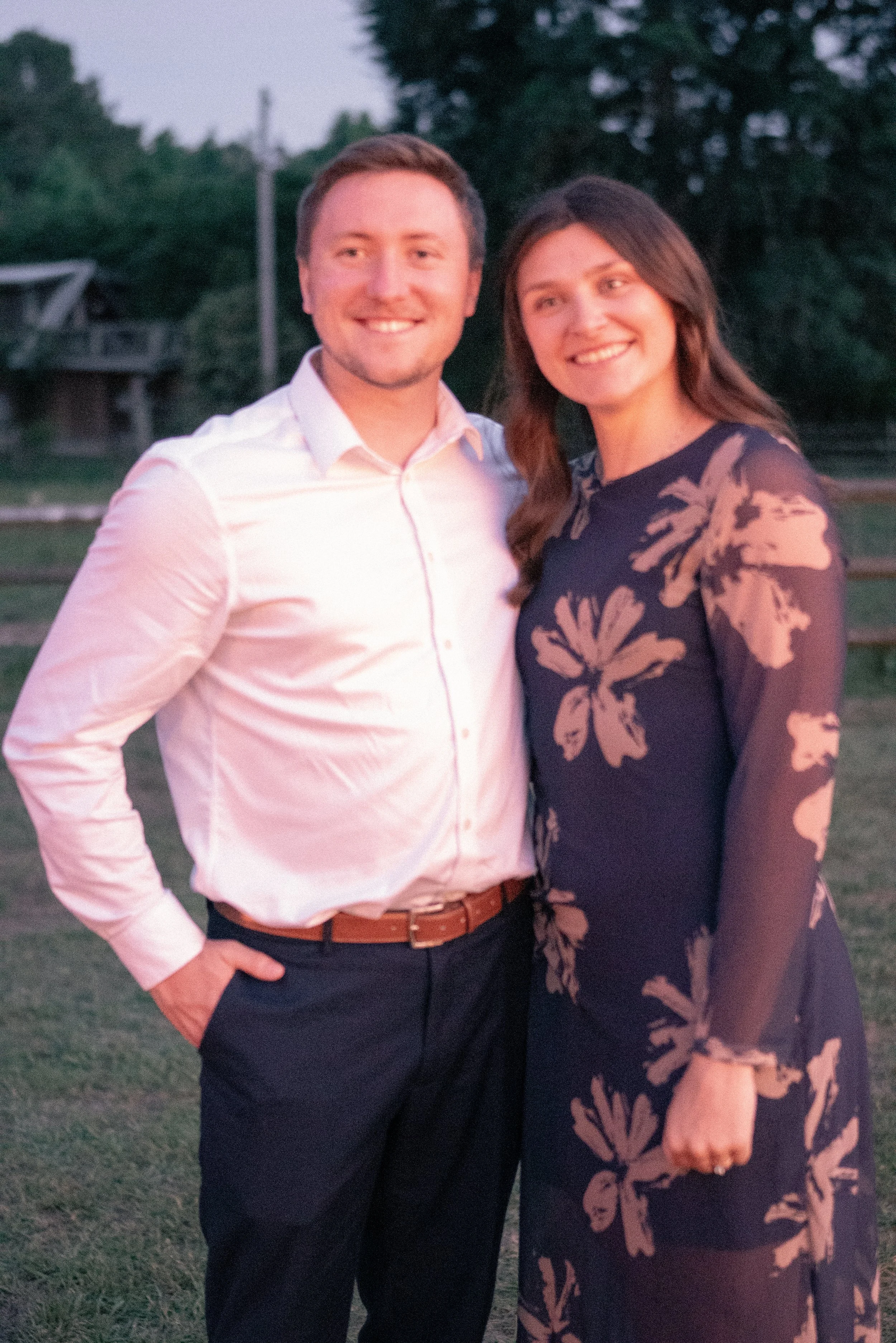 A man and woman standing close together outdoors during dusk, smiling at the camera, with trees and a house in the background.