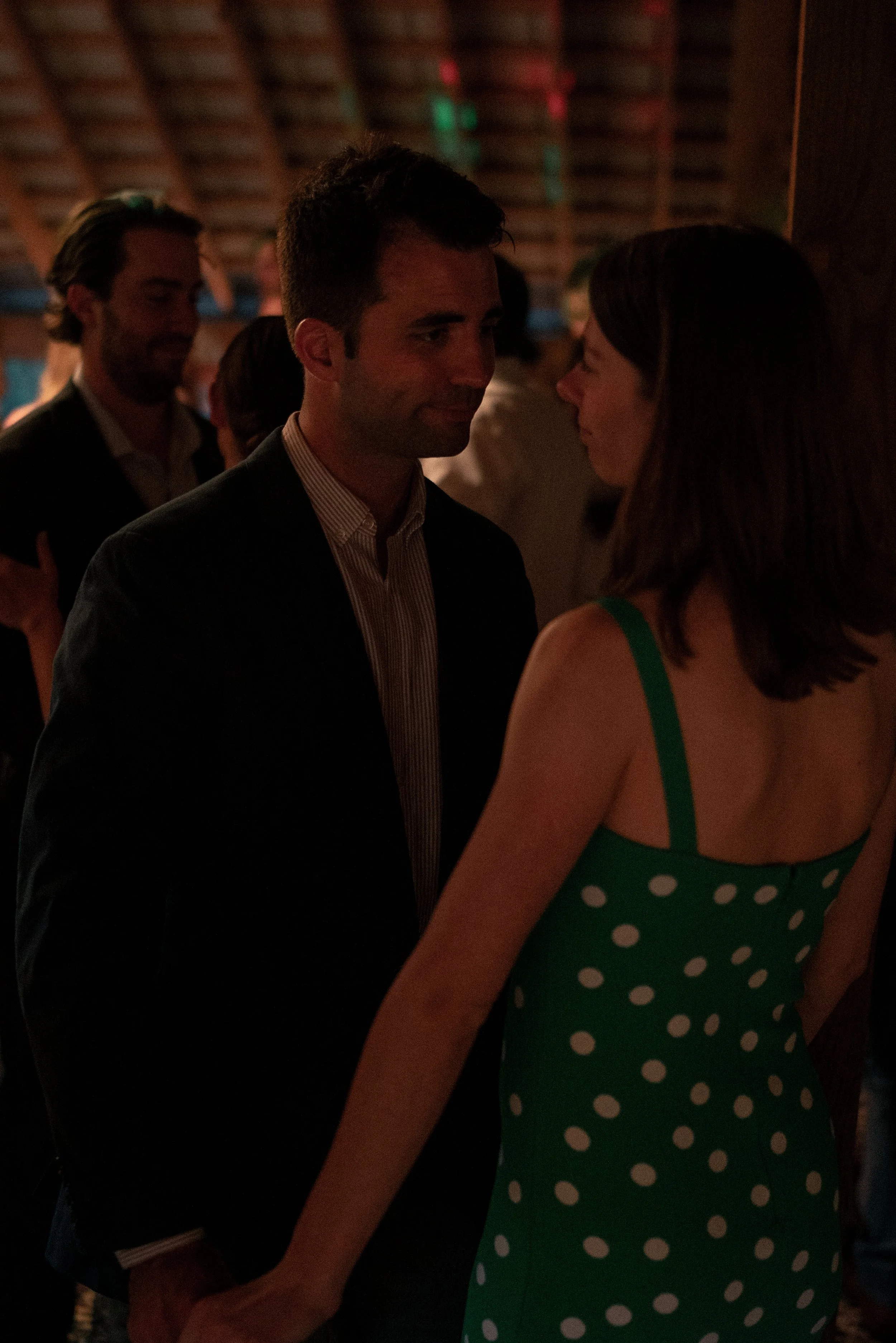 A man and woman facing each other and holding hands in a dimly lit indoor setting, possibly at a party or event.