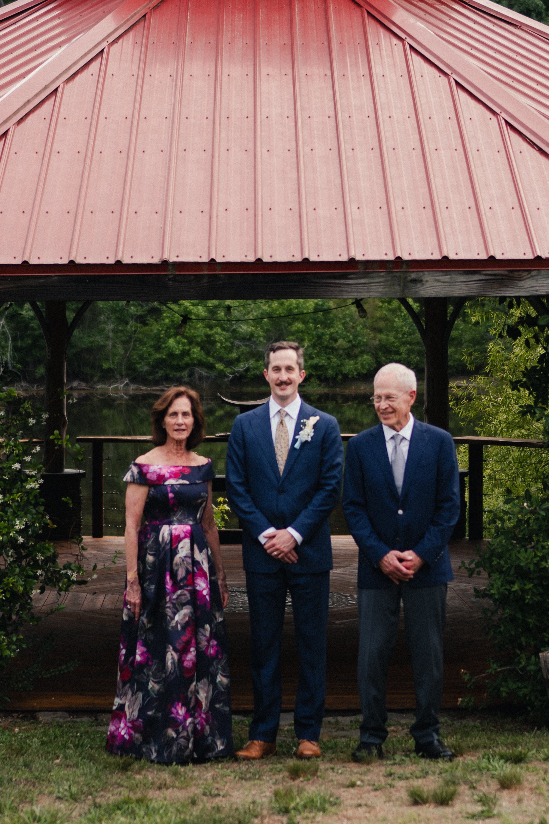 Three people standing outside on a wooden deck with a red metal roofed structure in the background. The group includes an older woman in a floral dress, a young man in a suit with a boutonniere, and an older man in a suit. There is greenery and water