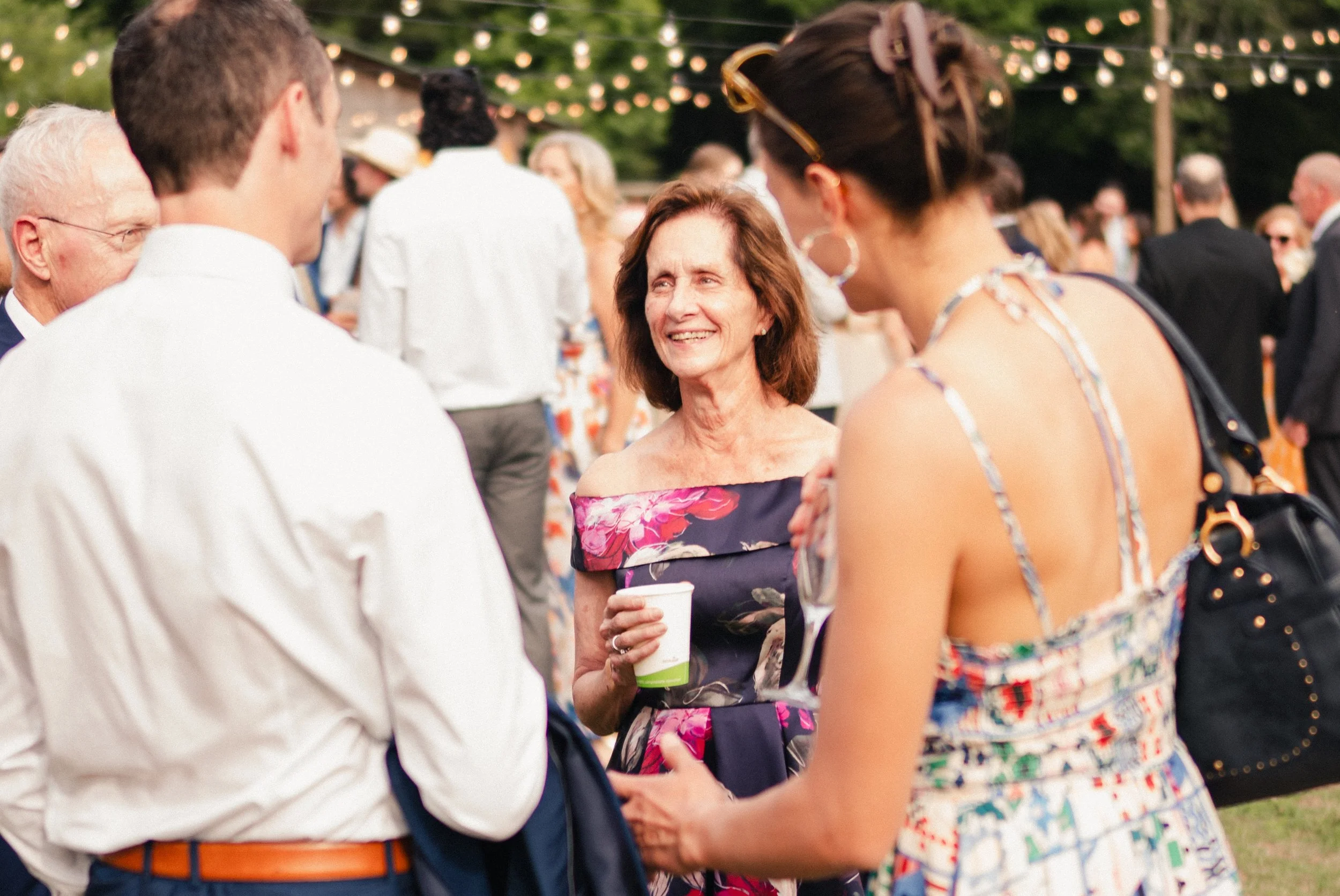 A woman with shoulder-length brown hair and a floral off-shoulder dress smiling as she interacts with a group of people at an outdoor gathering, with string lights hanging above and other guests chatting in the background.