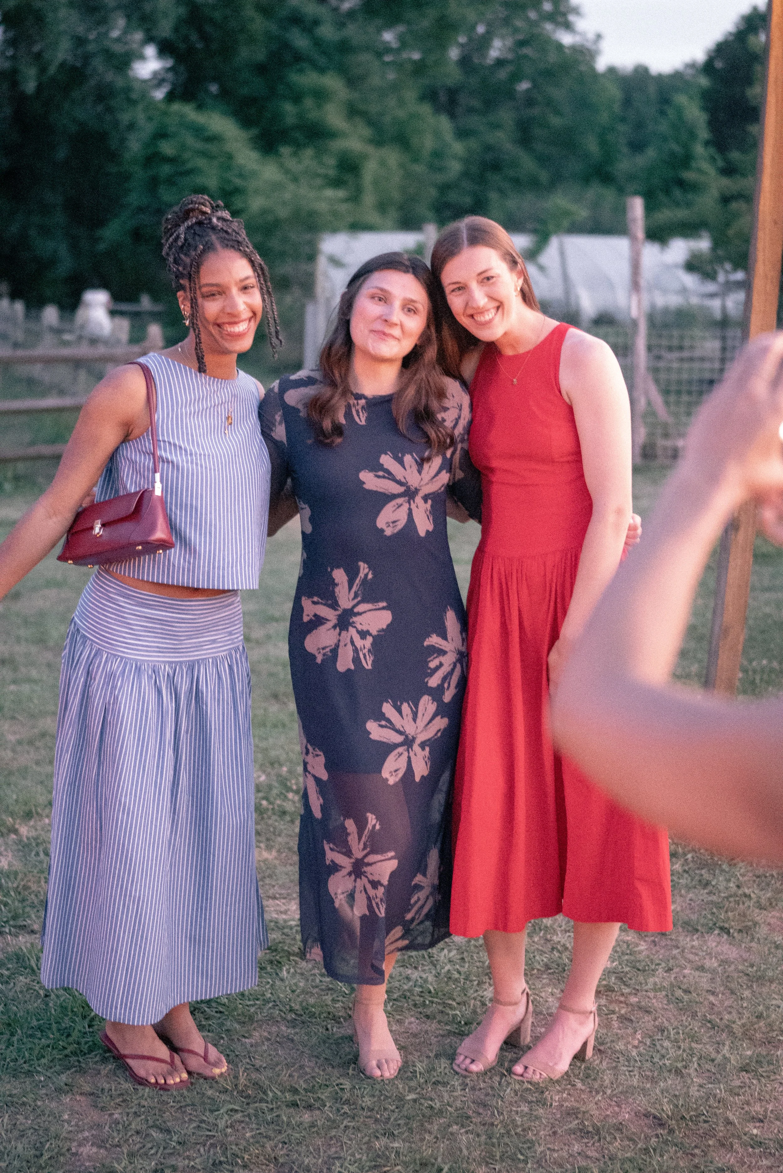 Three women dressed in summer dresses, standing together outdoors at sunset, smiling and taking a picture with a camera. Green trees and a wooden fence are in the background.