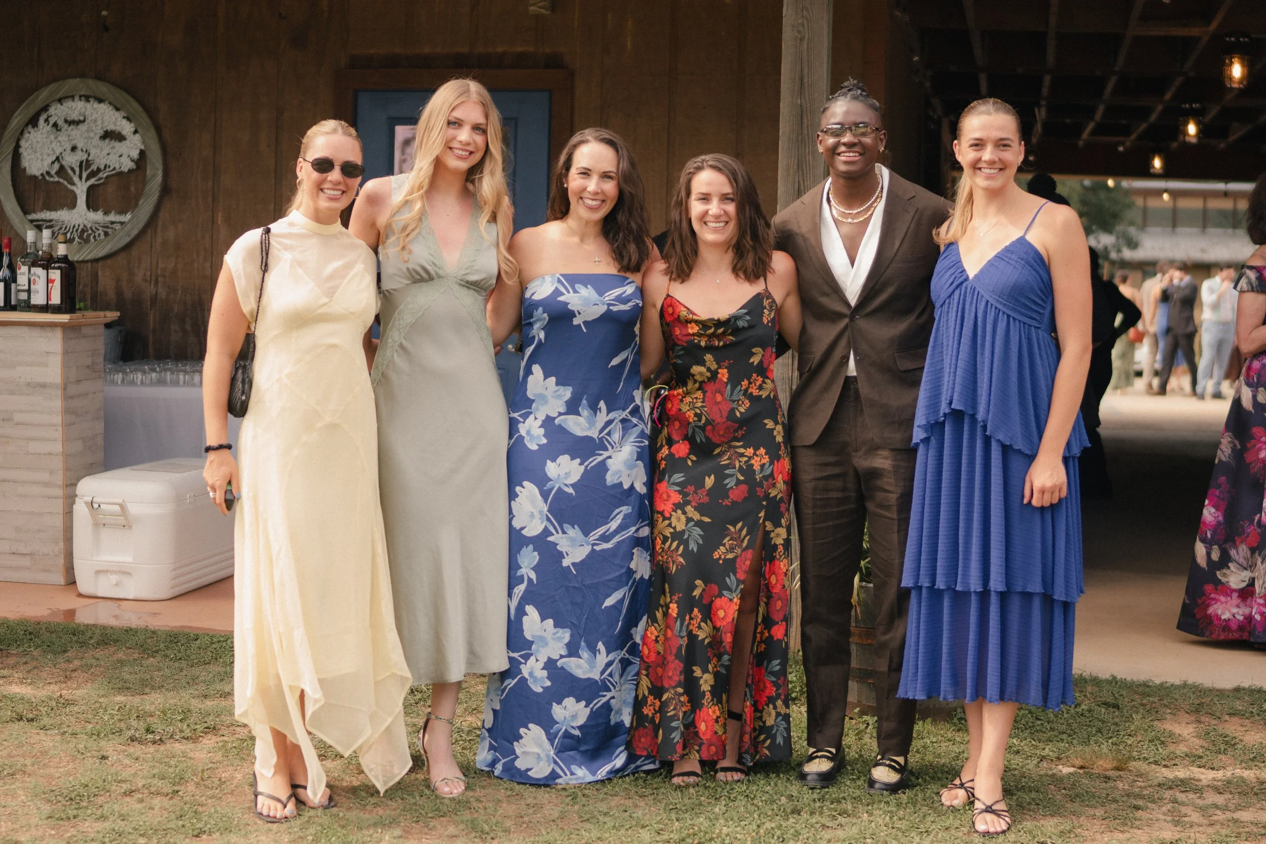 Group of six women standing together outdoors at a social gathering, smiling at the camera, dressed in colorful summer dresses, with some wearing sunglasses and jewelry.