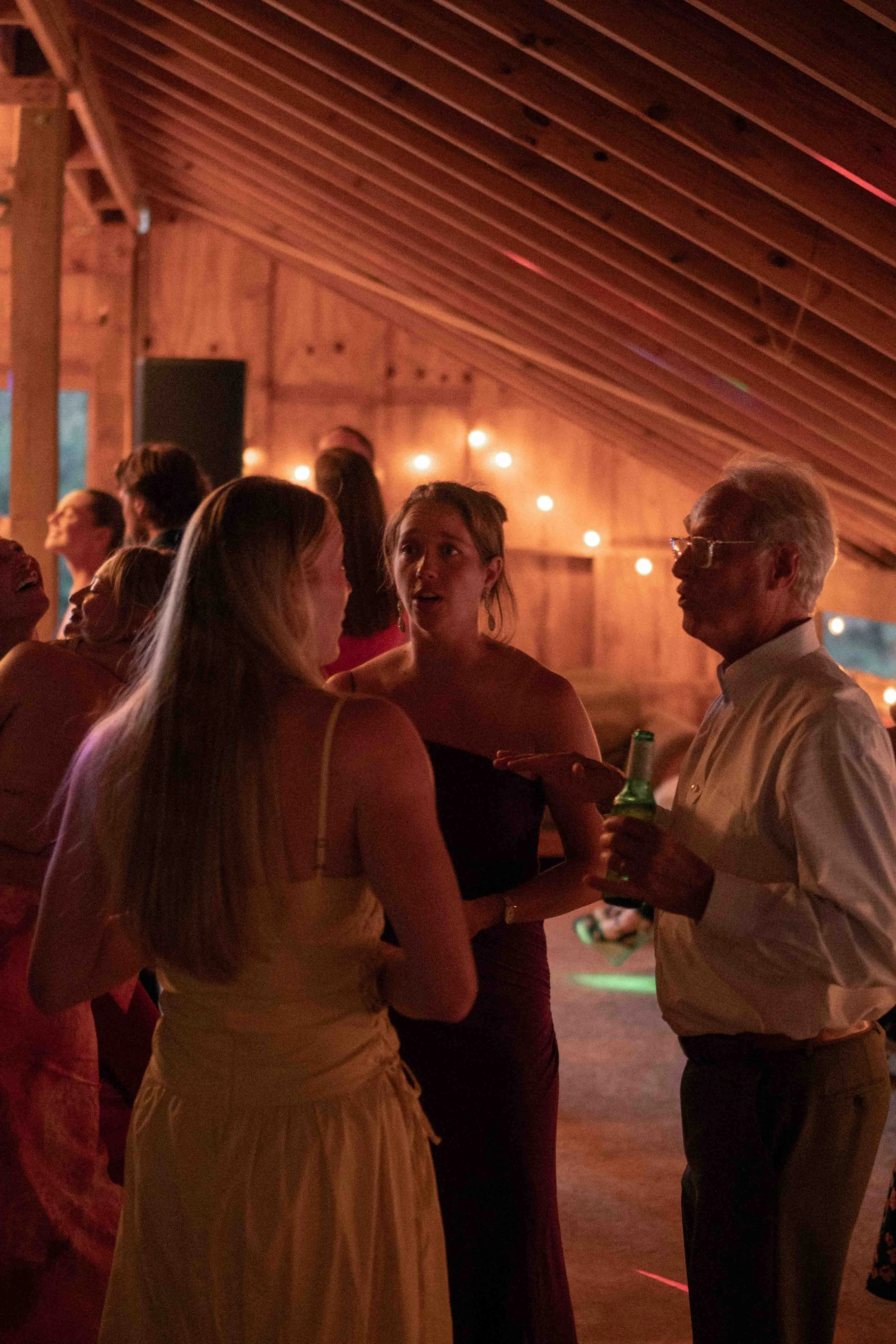 People gathered indoors at a social event, some holding drinks, engaging in conversations, with warm lighting and a wooden interior.
