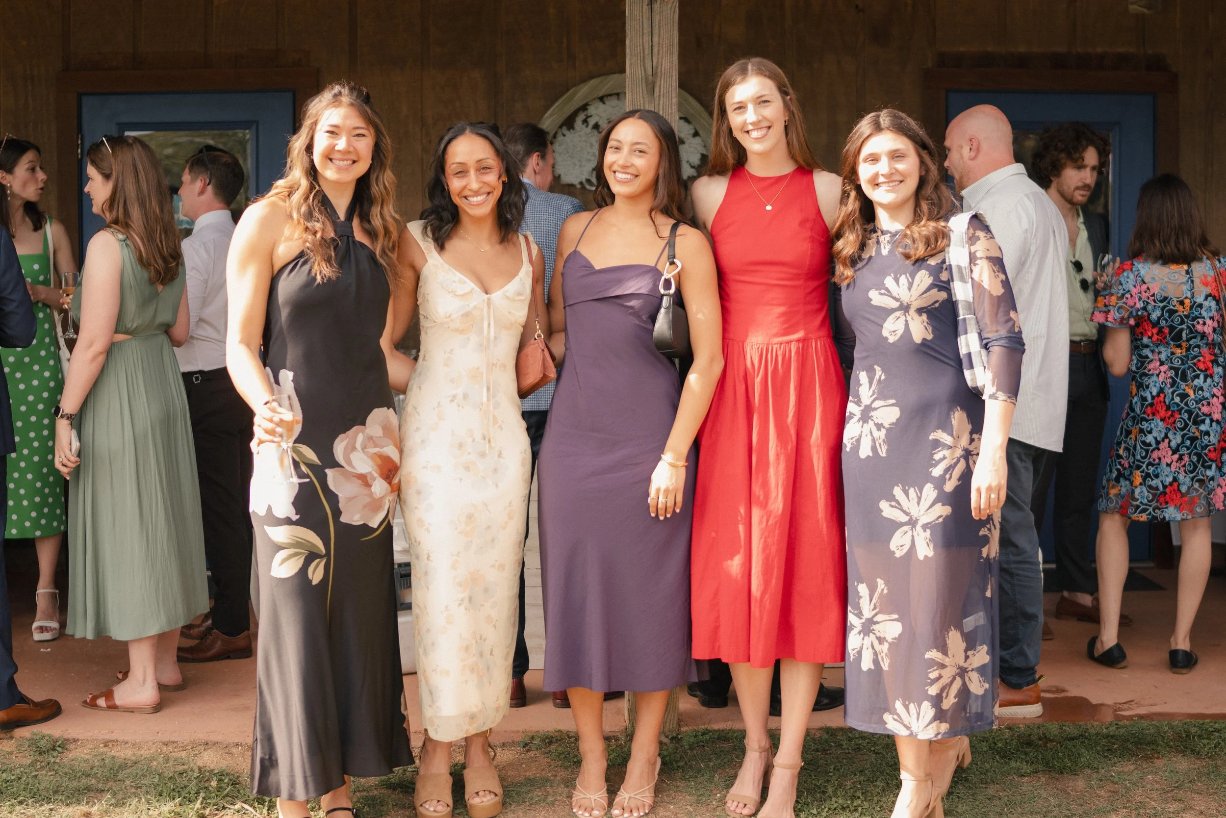Group of women in dresses smiling at a wedding or celebration, standing outdoors in front of a rustic wooden building.