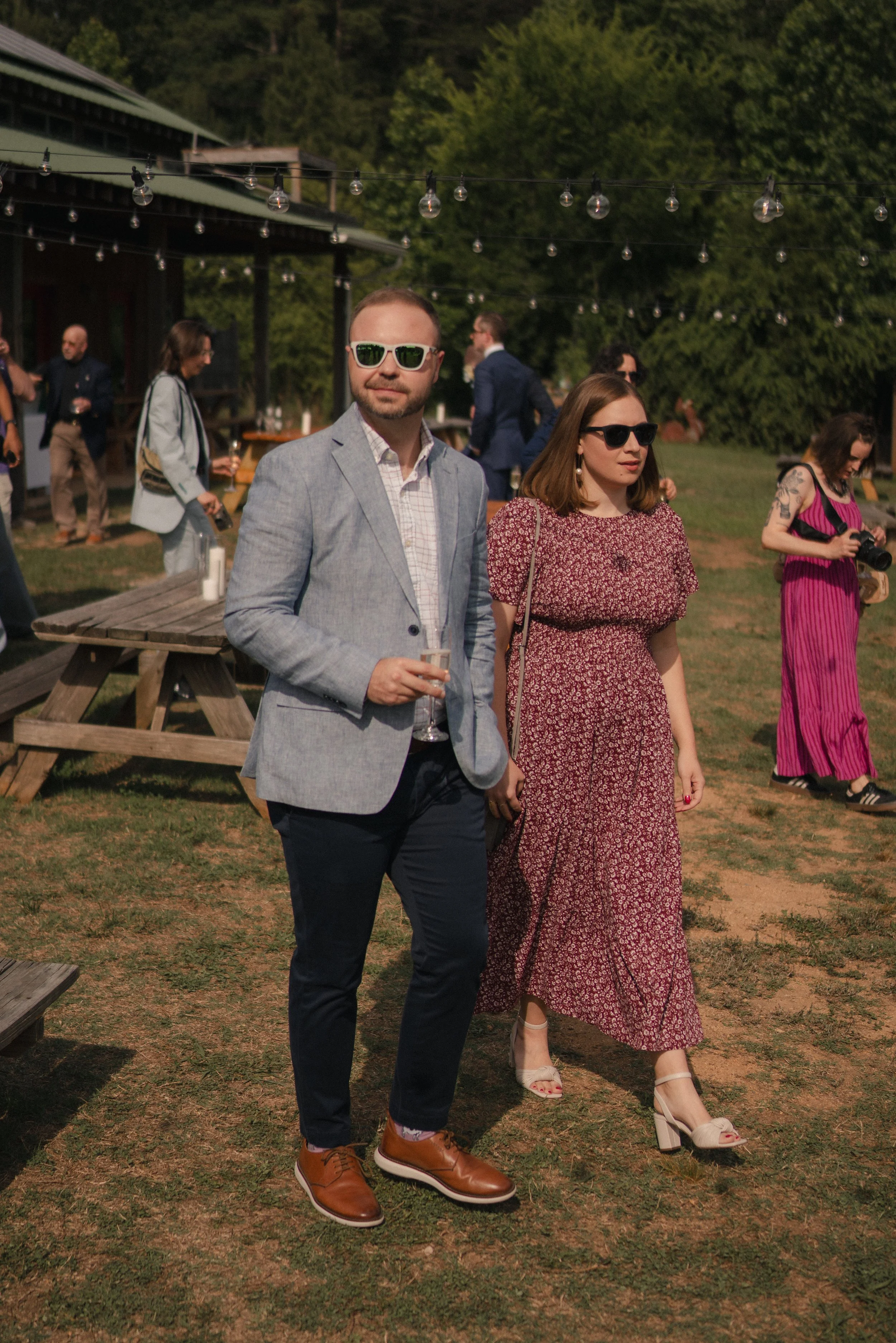 People dressed in formal attire at an outdoor event, with some holding drinks and others taking photos, under string lights with a green, wooded background.