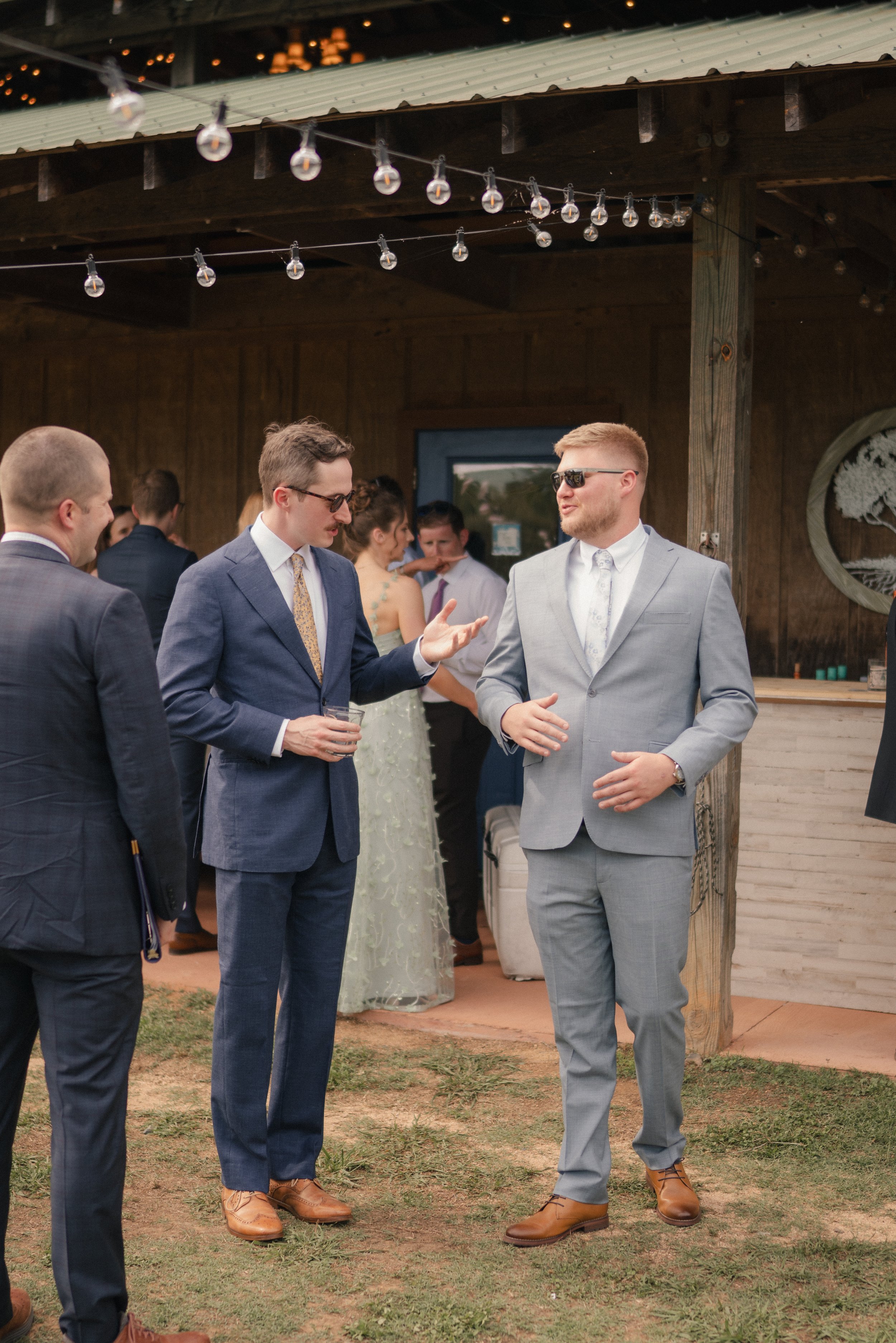 A group of men in suits engaged in conversation at an outdoor event during the evening. They are standing on a grassy area with a wooden building and string lights overhead.