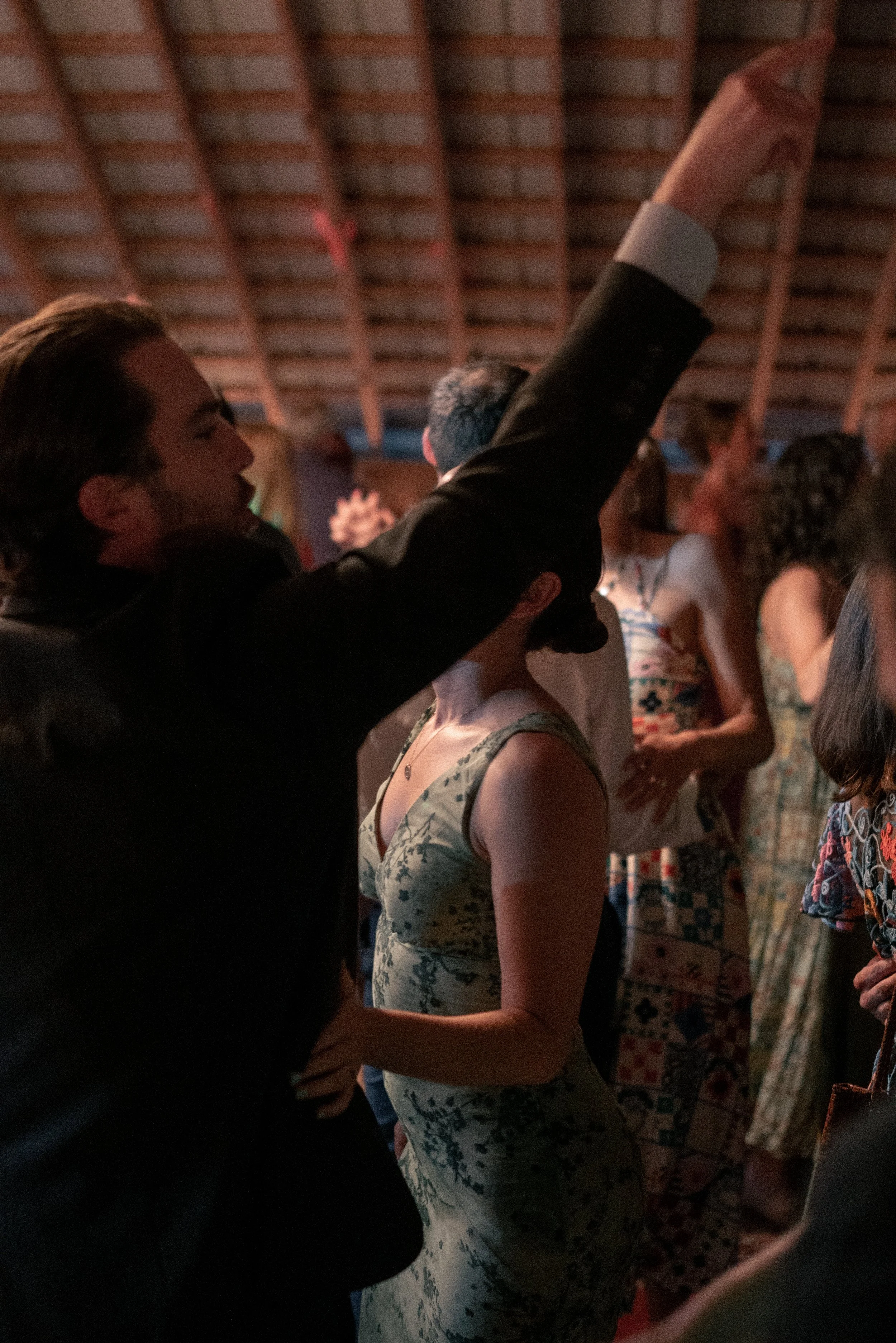 People gathered indoors, some with hands raised, possibly at a concert or event with a wooden ceiling visible.