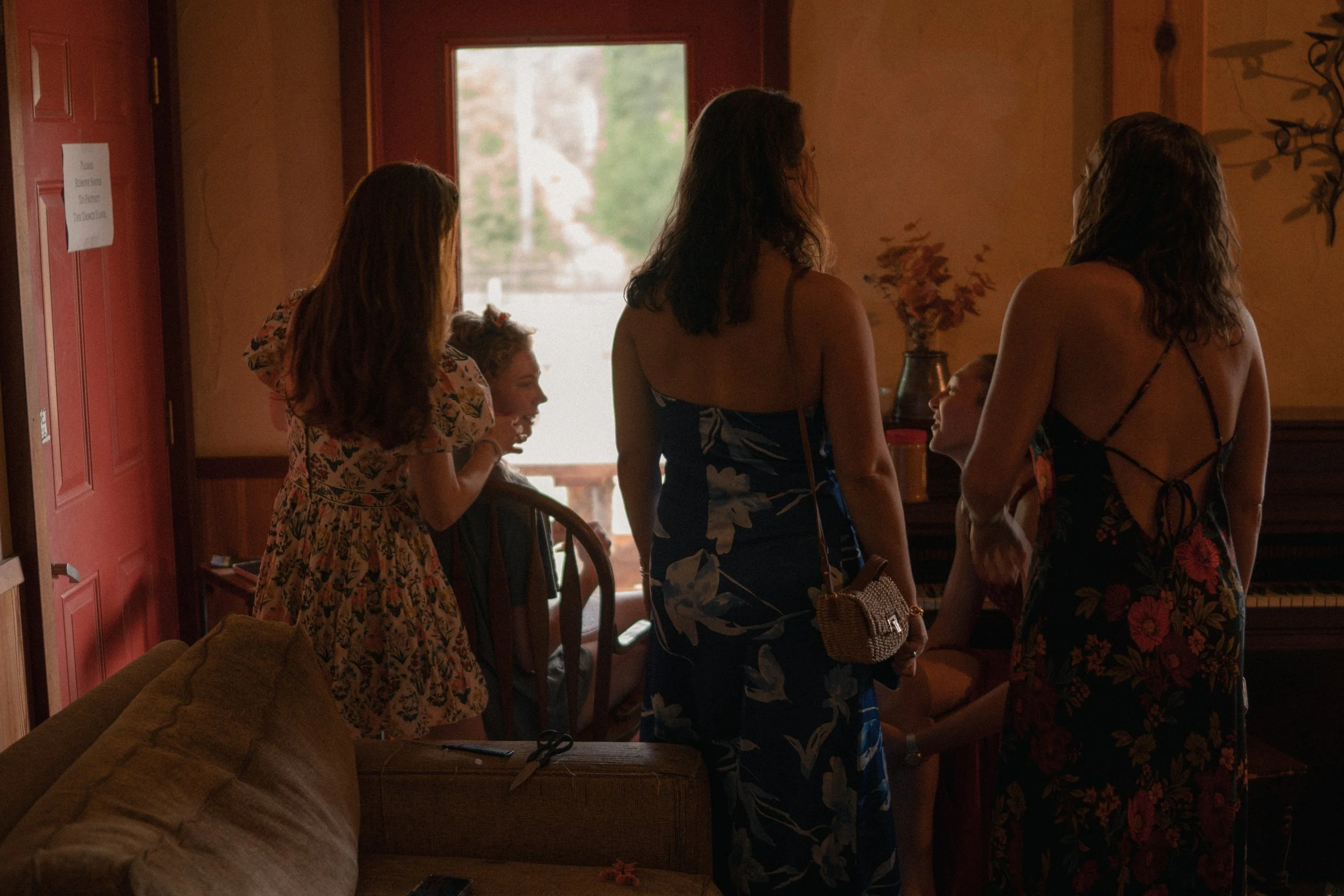 Group of women and a girl gathered indoors near a window, engaging in conversation, with visible floral dresses and a warm, cozy interior.