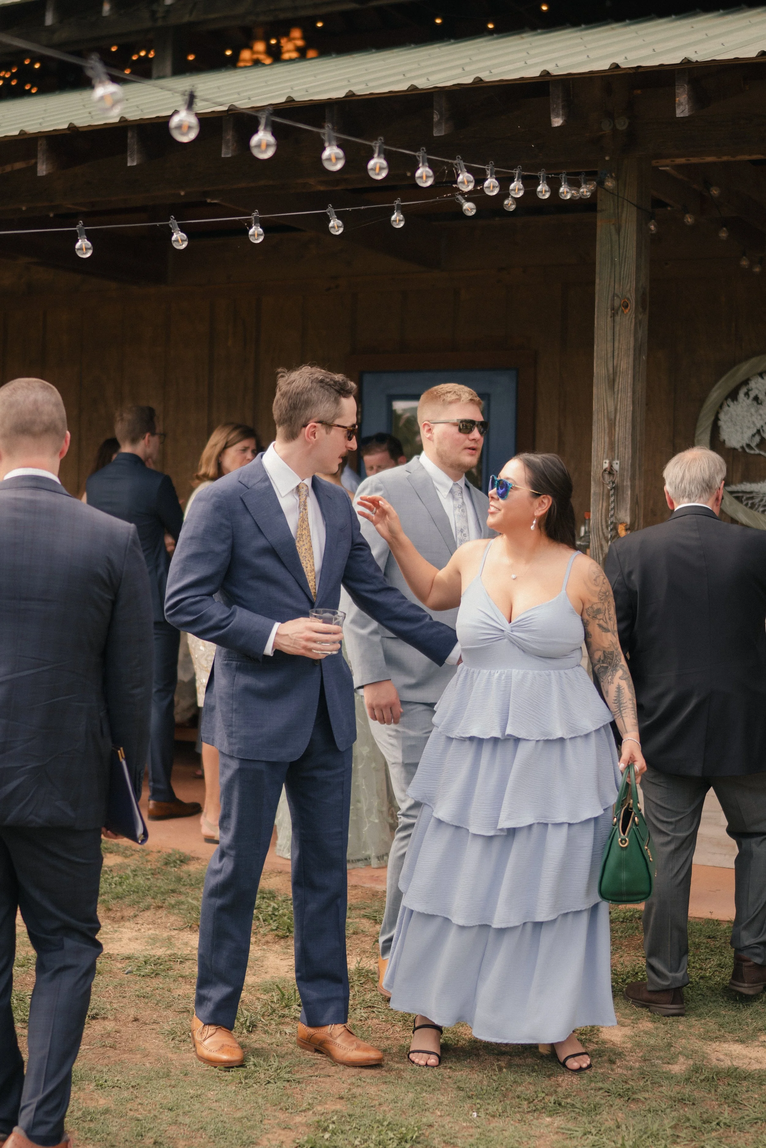 People attending an outdoor wedding reception, some wearing formal attire and sunglasses, under a string of lights with a rustic wooden backdrop.