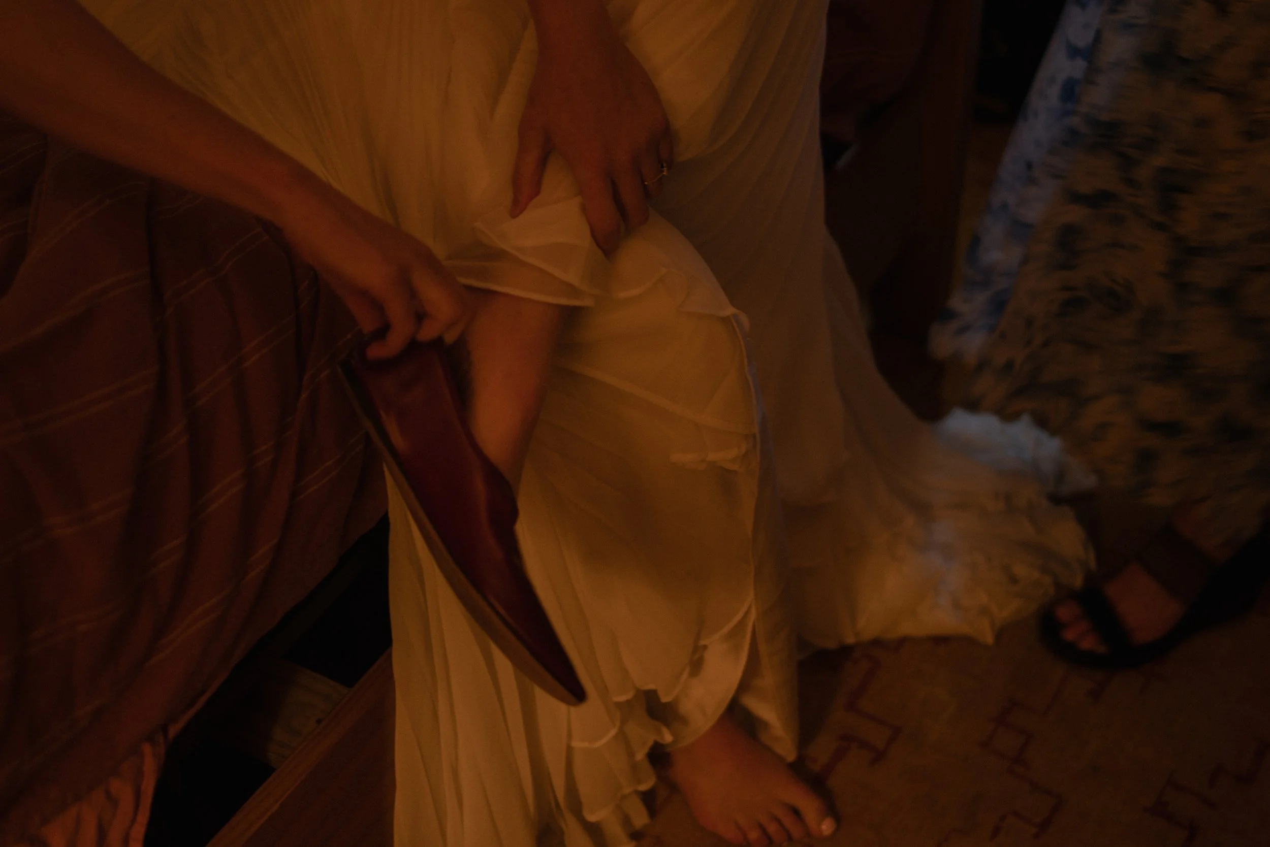 Close-up of a person's hand adjusting the ruffled lower part of a light-colored dress. The background shows part of a patterned carpet.