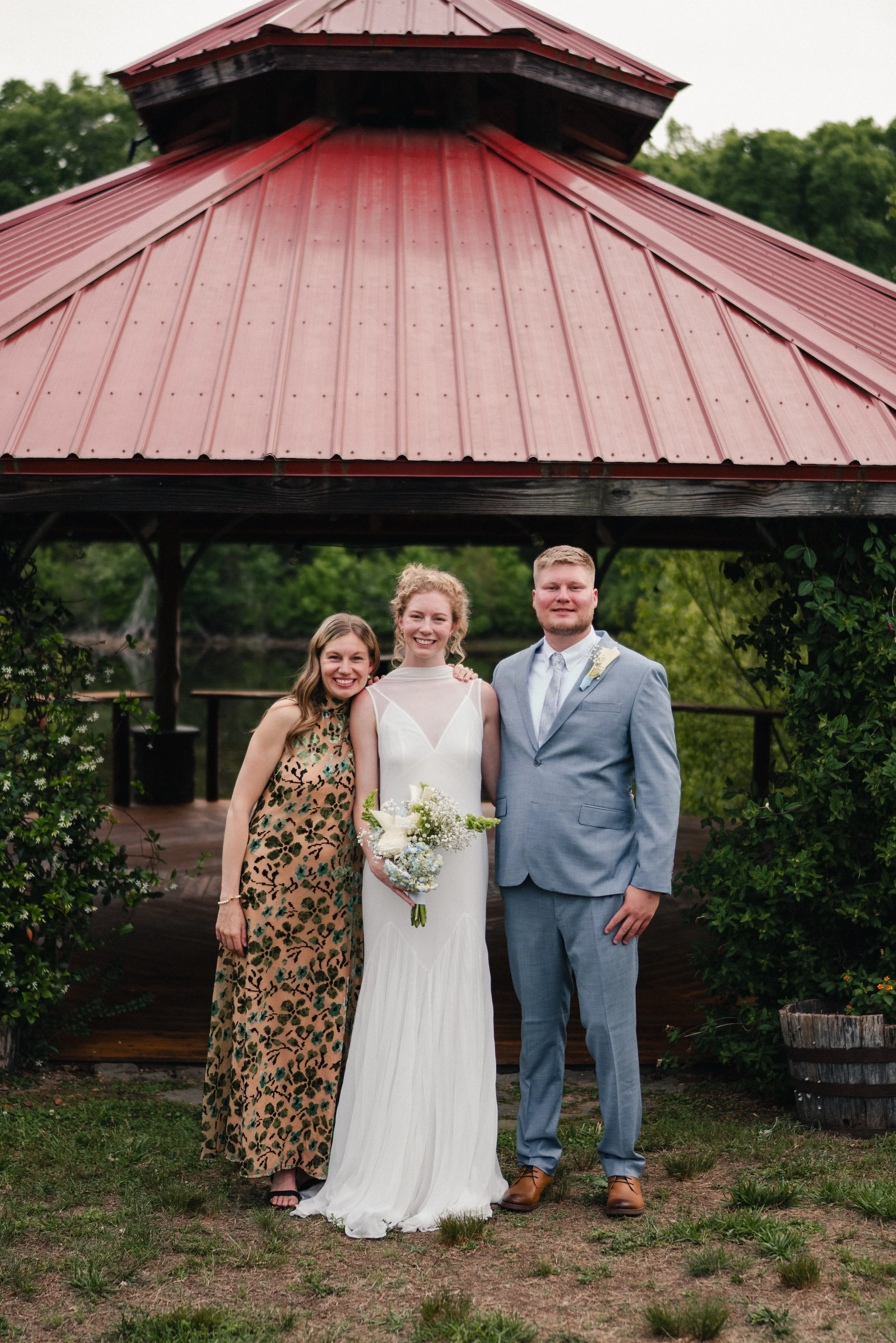Group of three people standing outdoors under a gazebo, smiling at the camera, with a woman wearing a wedding dress holding a bouquet of flowers.
