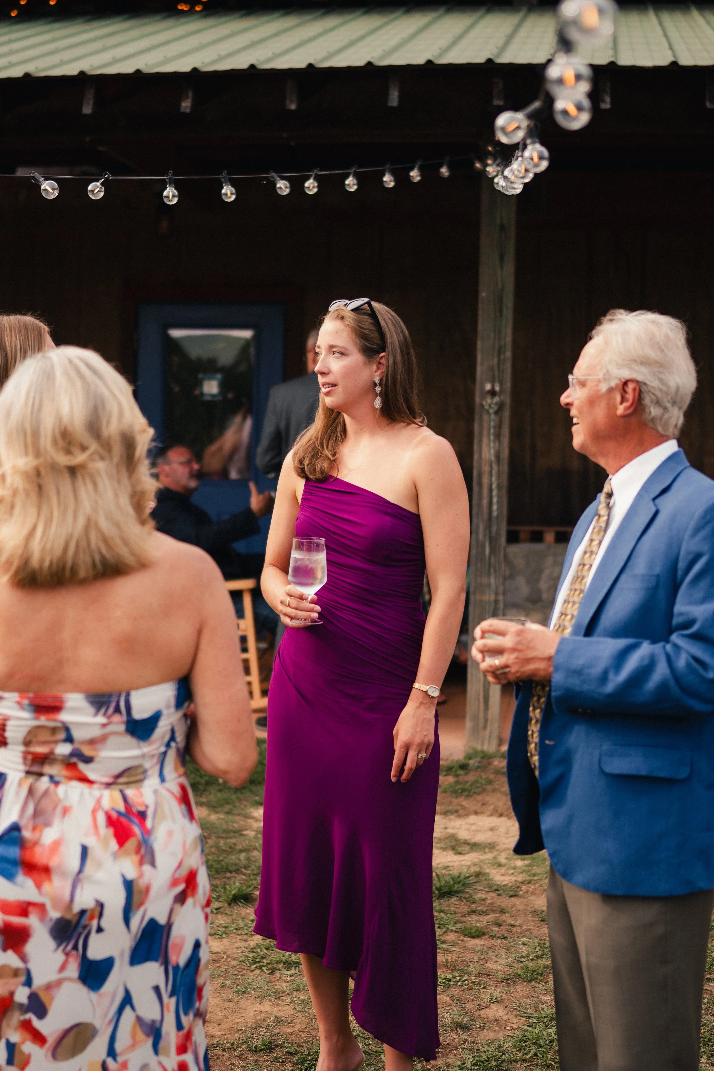 People socializing at an outdoor gathering under string lights, with a woman in a purple dress holding a glass of wine.