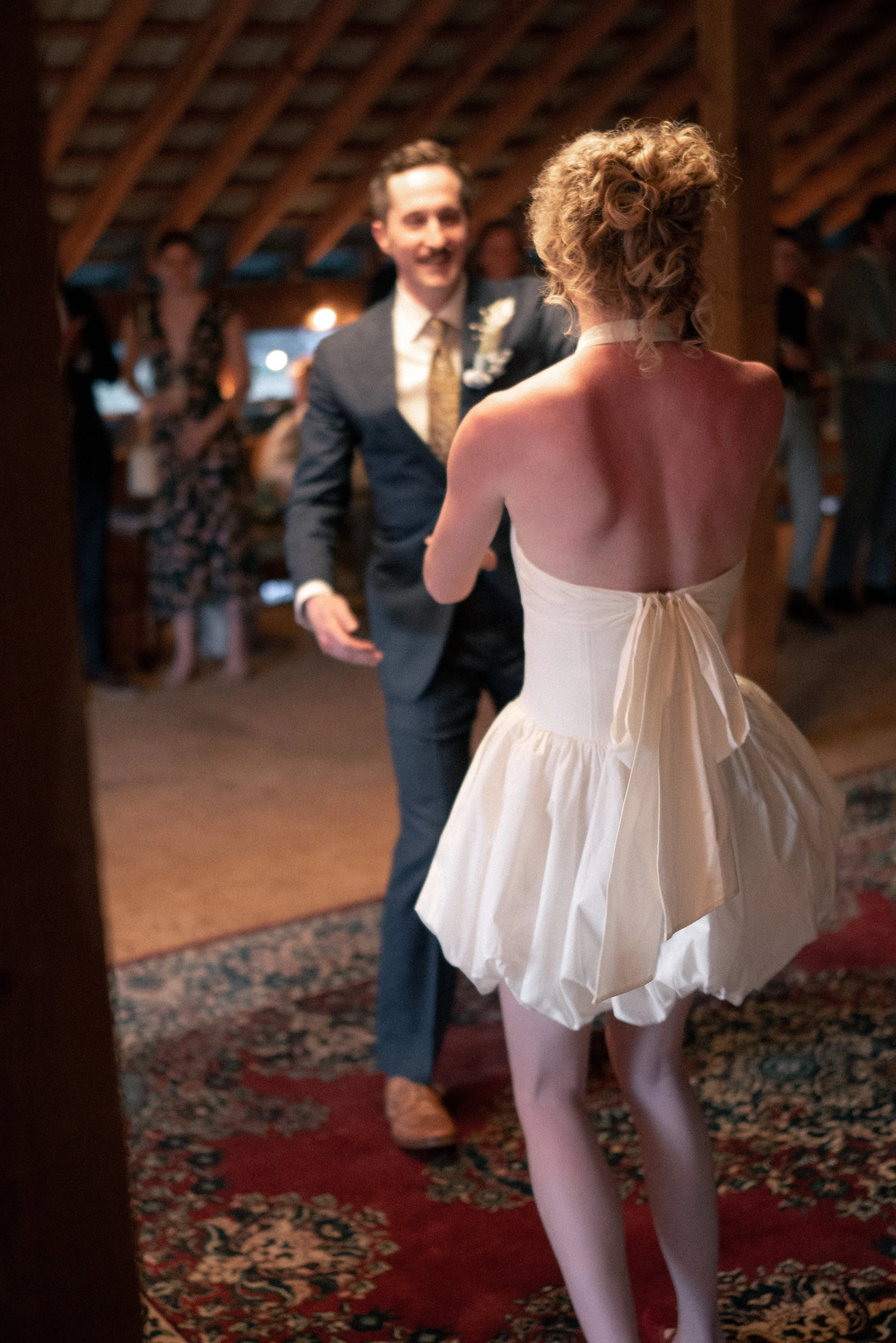 A wedding dance with a bride in a white strapless dress and a groom in a dark suit, both smiling, surrounded by guests in a rustic venue with wooden beams and a patterned rug.