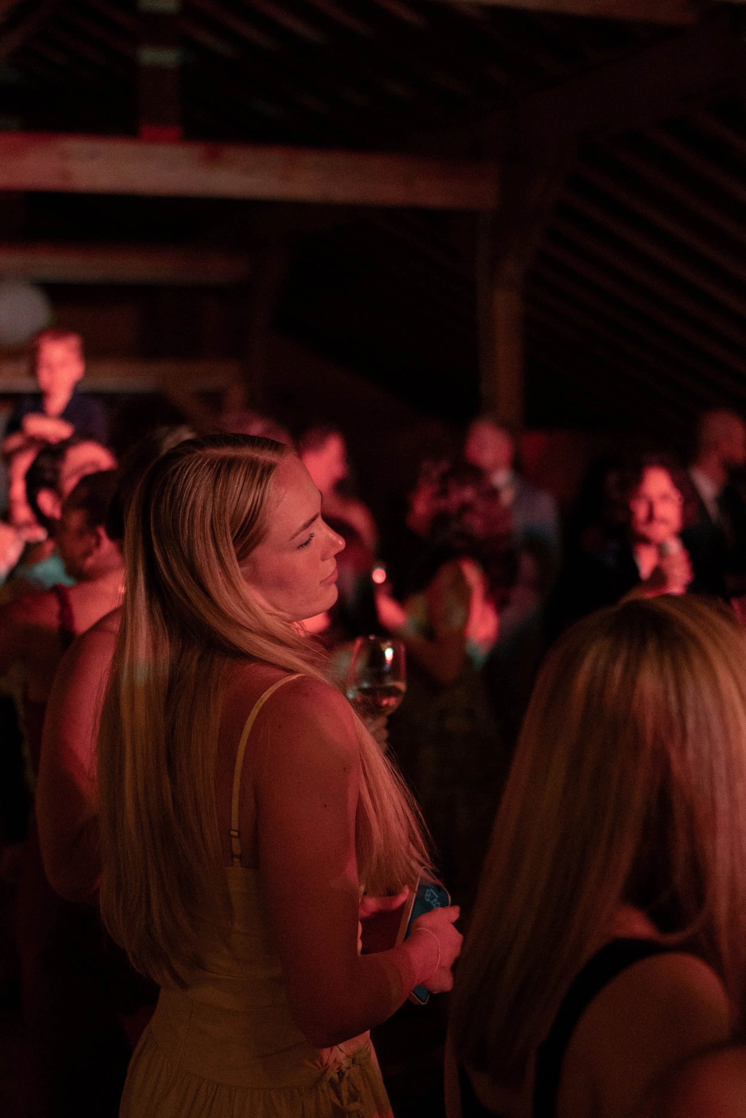 A woman with long blonde hair in a yellow dress holding a glass of wine at a crowded event with dim, red lighting and wooden ceiling.