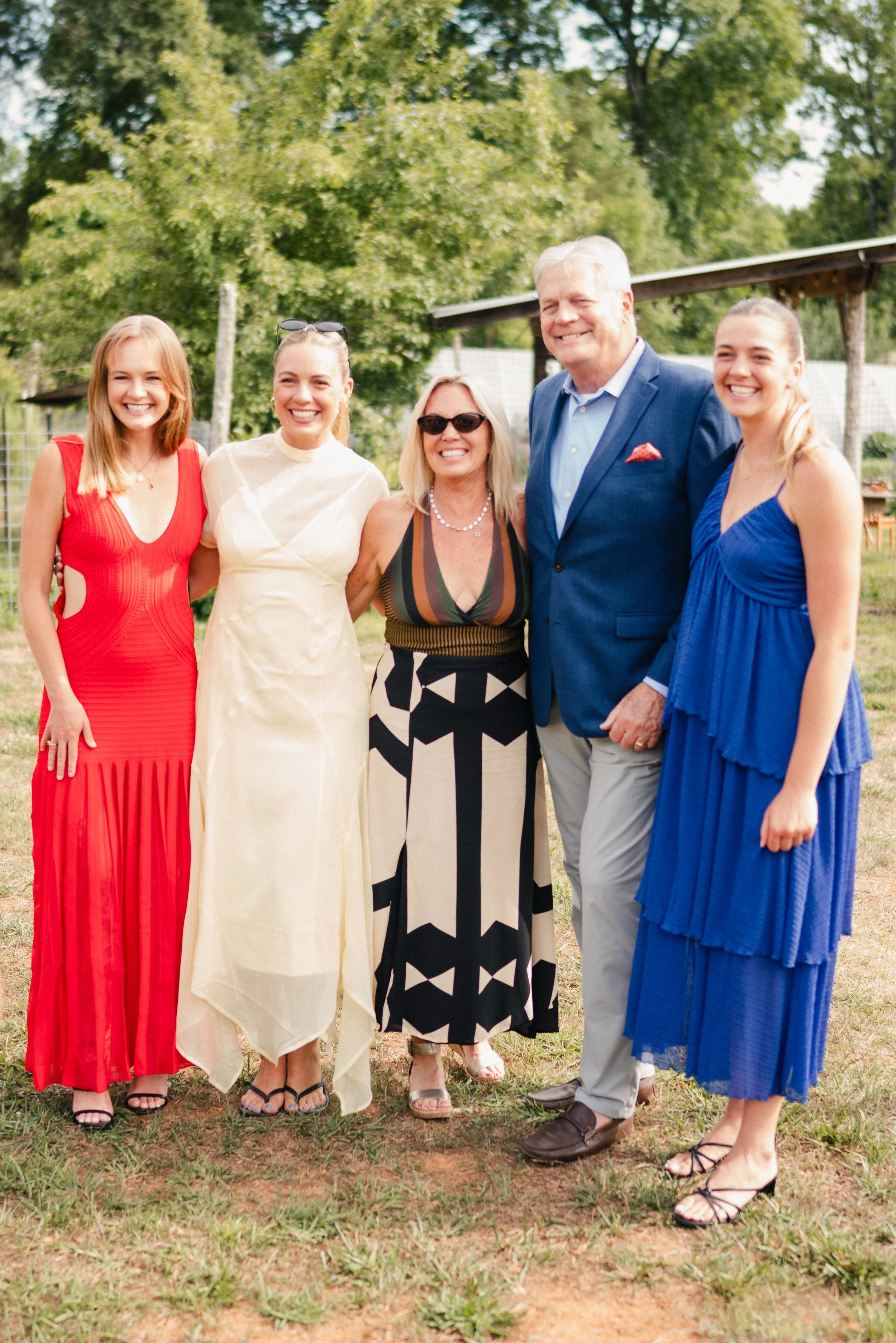 Group of five women and one man standing outdoors during daytime, smiling for a photo. They are dressed in colorful summer dresses and a suit. Behind them are green trees and a wooden structure.