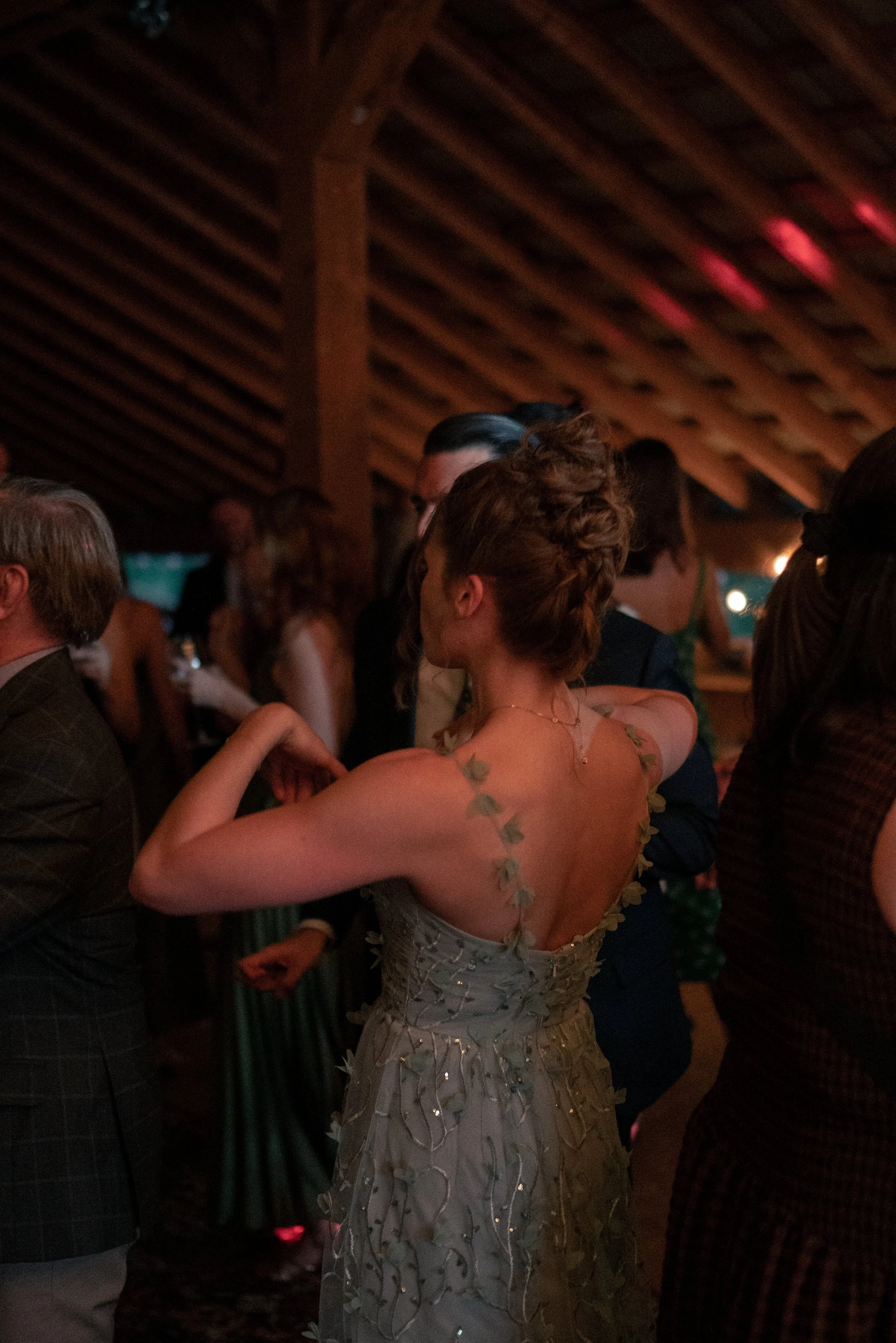 People dancing at a wedding reception, with a woman in a floral gown in the foreground and a wooden ceiling in the background.