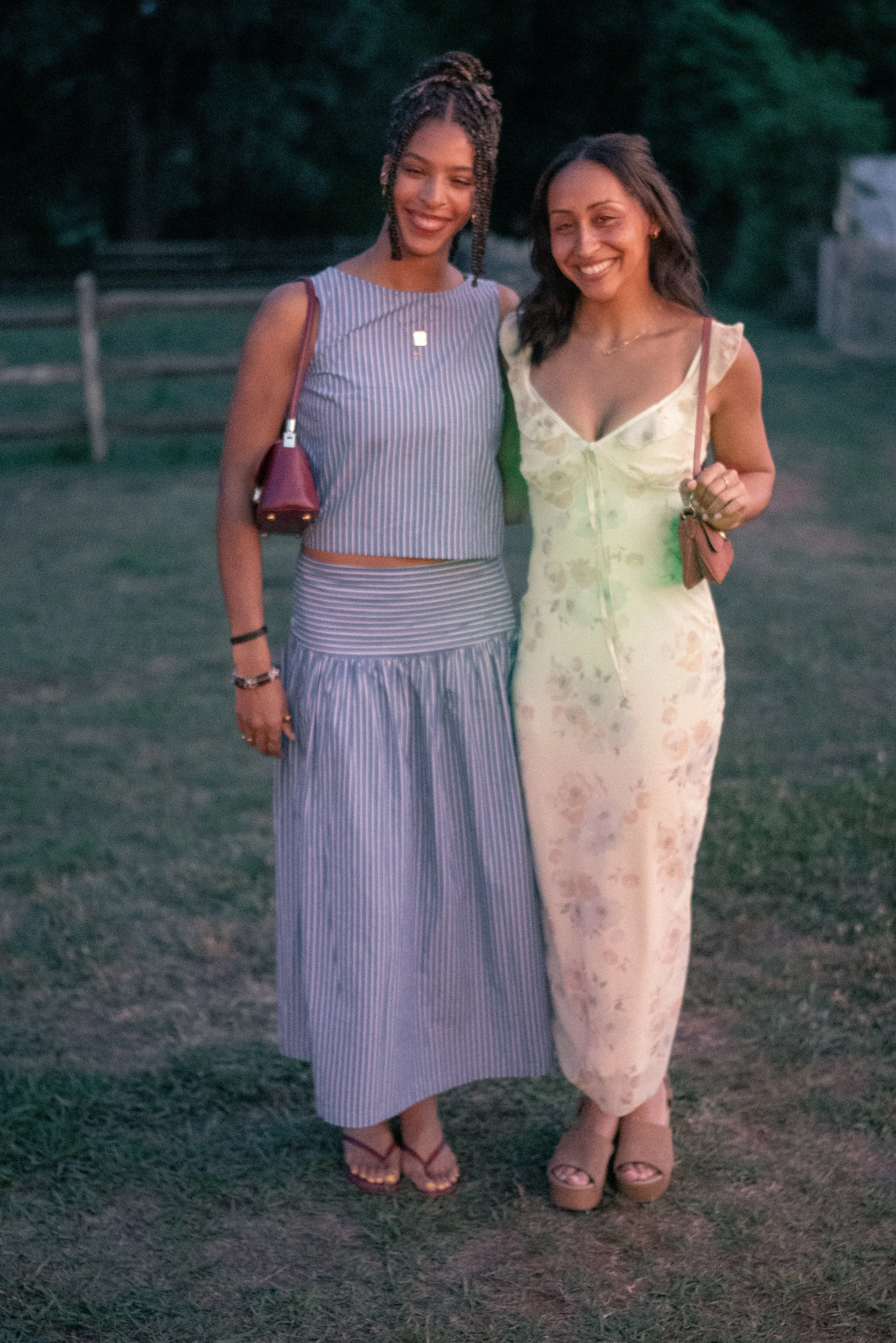 Two women standing outdoors on grass, smiling, with trees and a fence in the background, during dusk or evening.