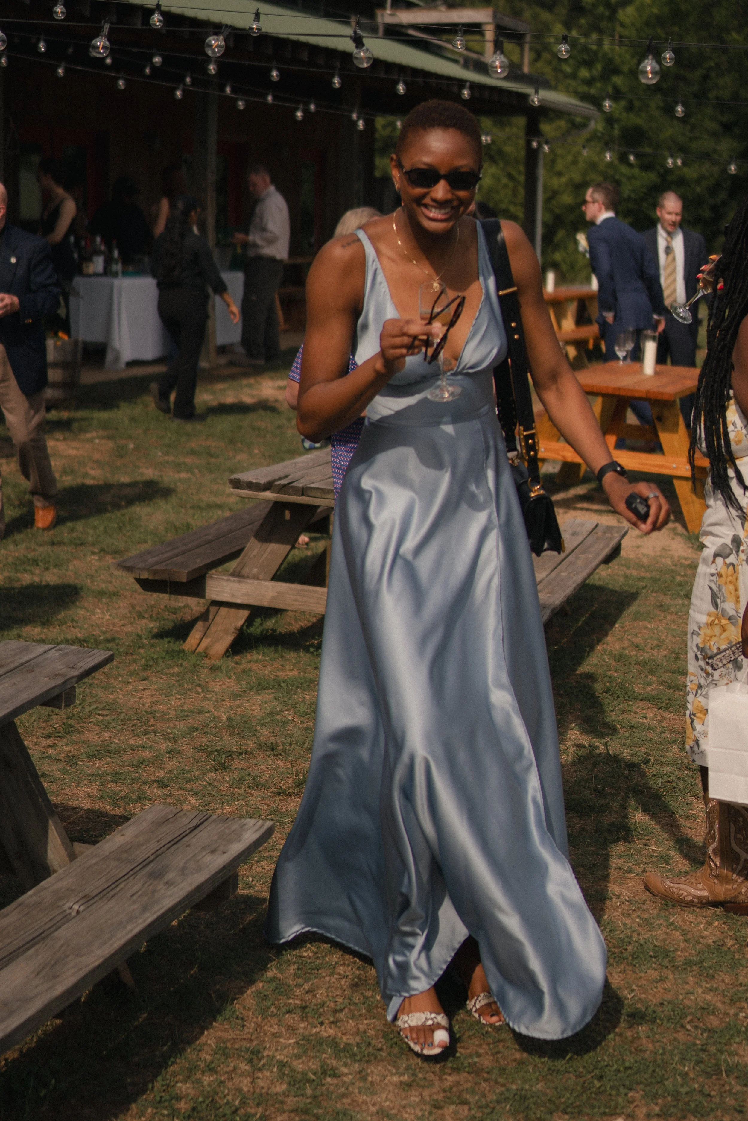 A woman in a shiny blue dress holding glasses, smiling, standing outdoors at a social gathering with people in the background and string lights overhead.
