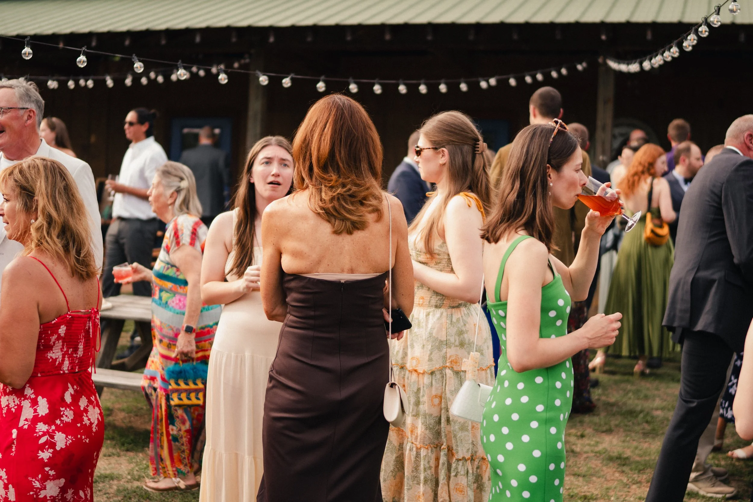 People socializing at an outdoor gathering, some wearing colorful dresses and holding drinks, under string lights with a wooden structure in the background.