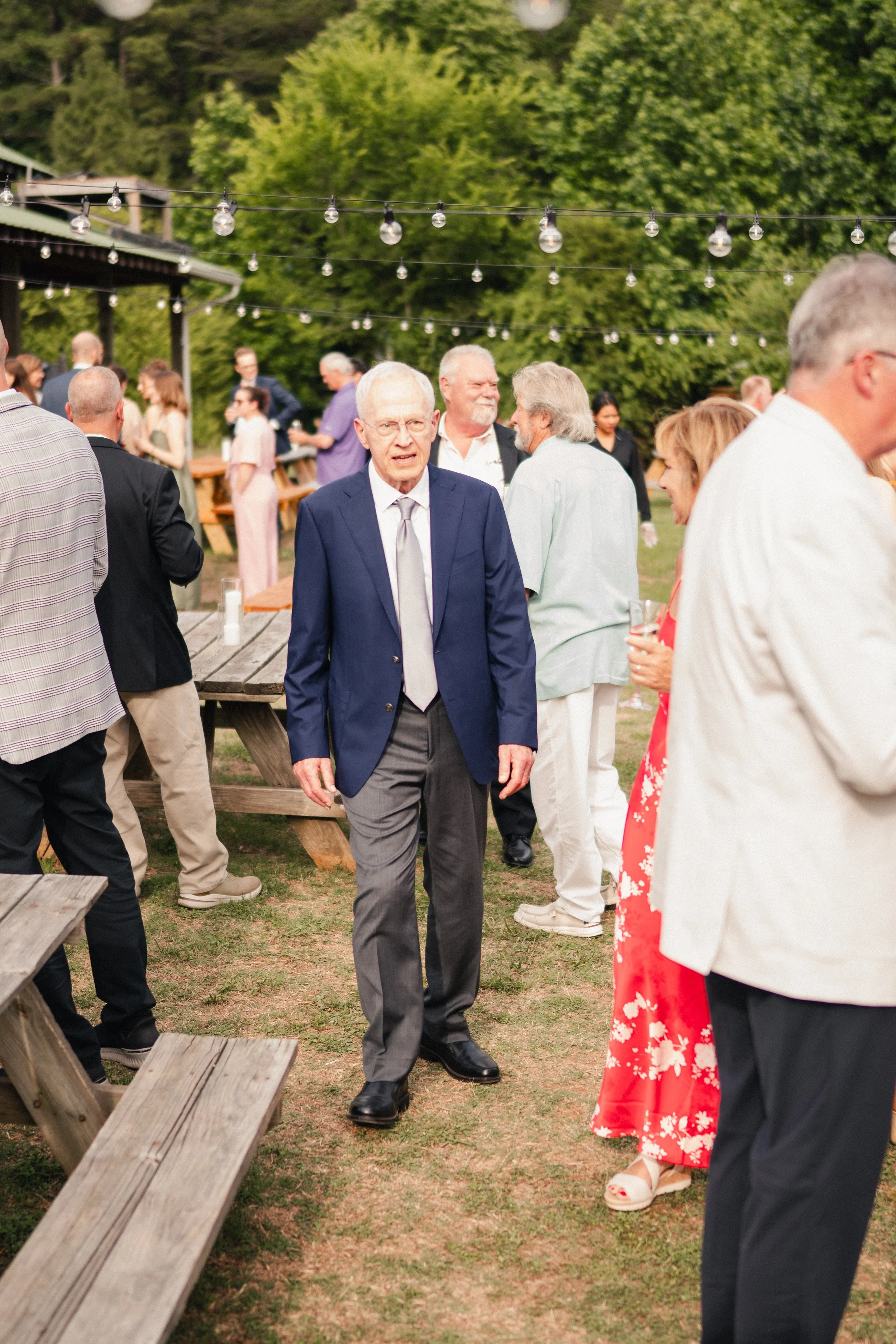 People gathered outdoors in formal attire, socializing at what appears to be a celebration or event, with string lights hanging above and a green, wooded background.