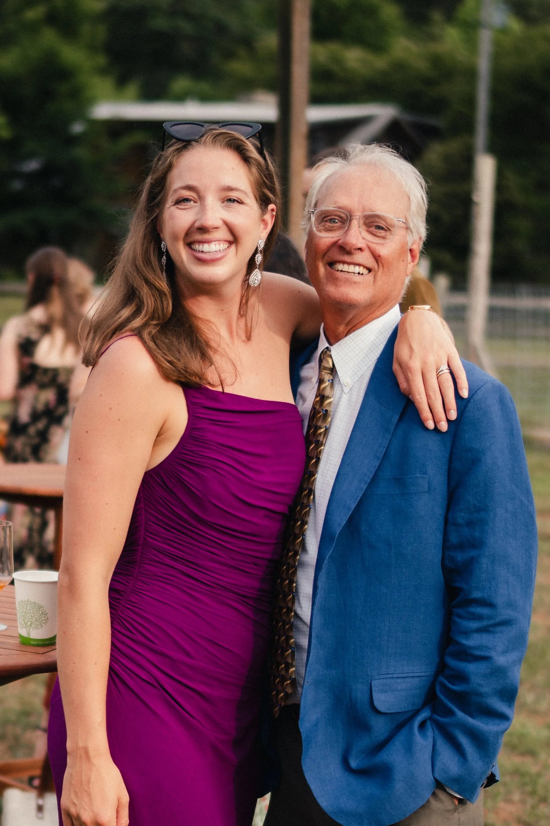 A young woman in a purple dress and an older man in a blue suit are smiling and posing together outdoors at a social event.
