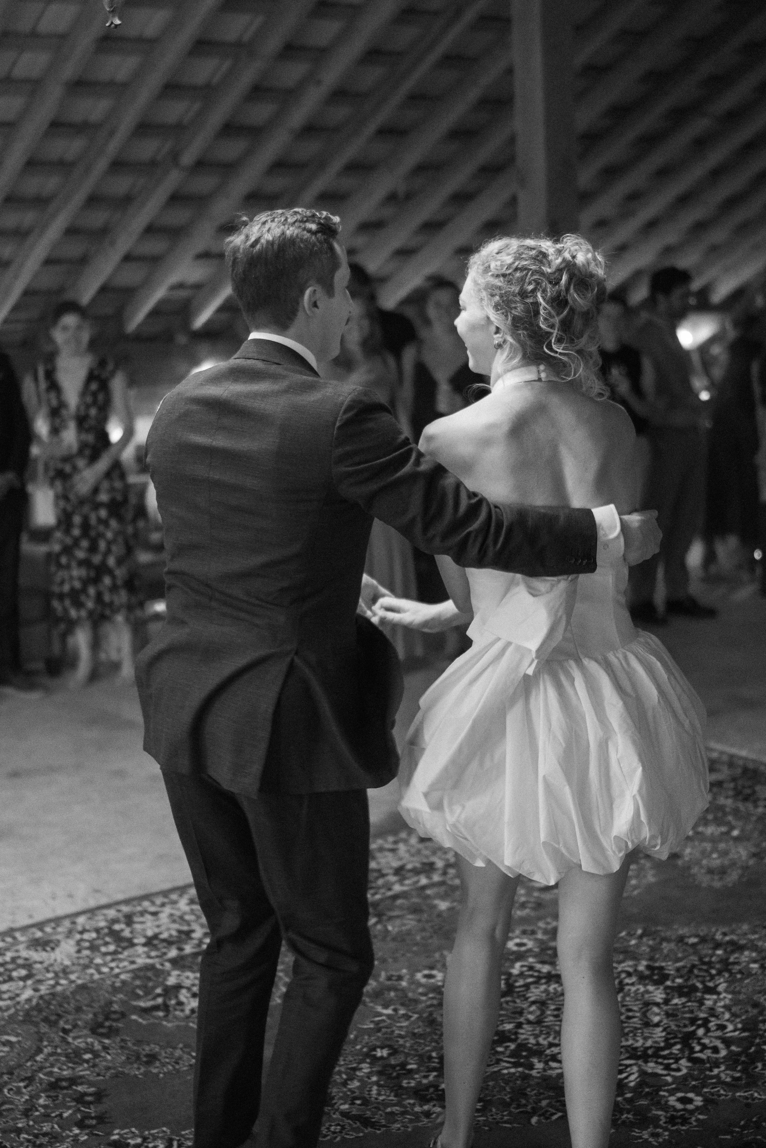 A couple dancing at a formal event, with the woman wearing a strapless white wedding dress and the man in a dark suit. They are holding hands and facing each other in a warmly lit venue with other guests in the background.