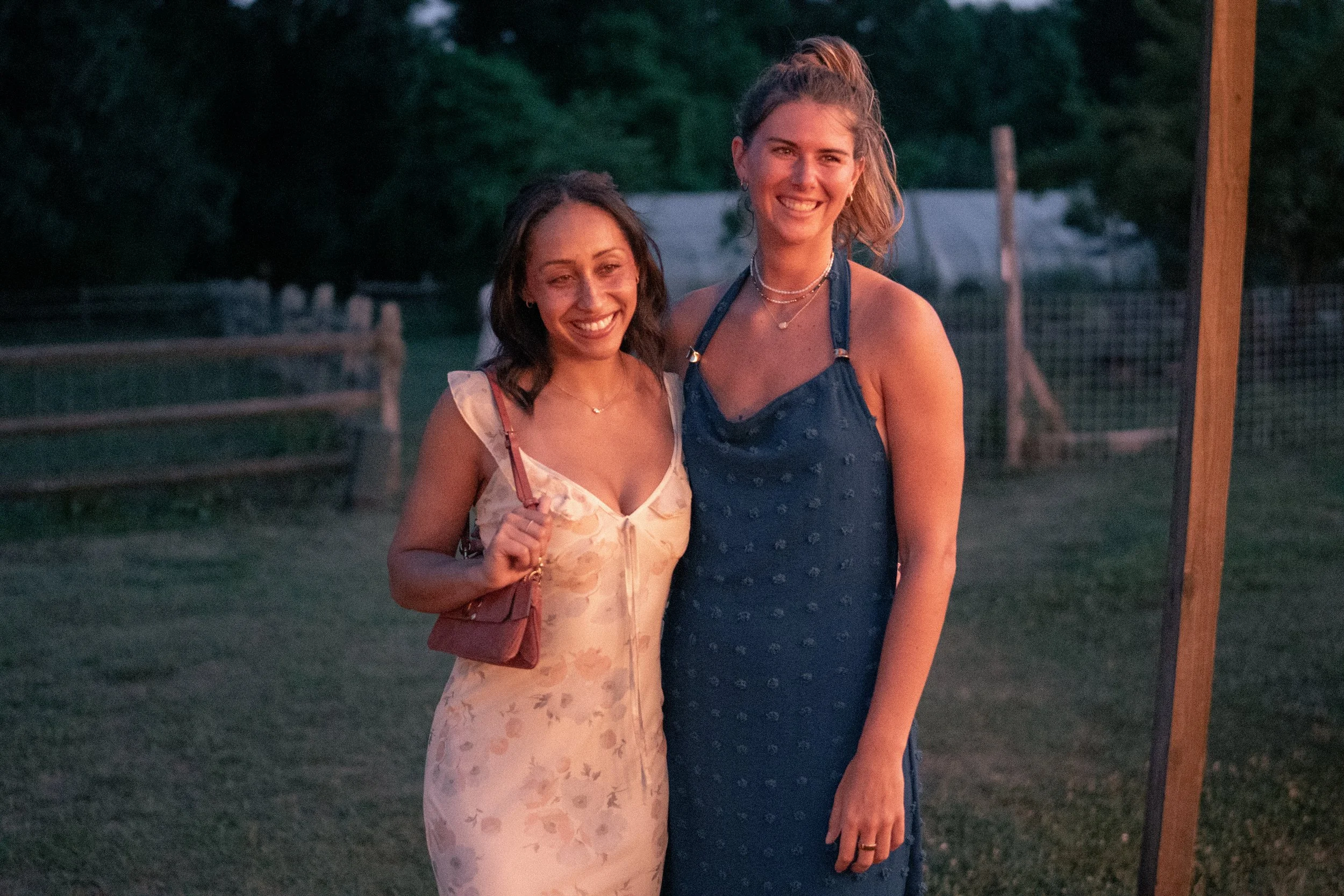 Two women standing outdoors at sunset, smiling, with trees and a wooden fence in the background. The woman on the left has dark hair and is wearing a light-colored dress, carrying a small purse. The woman on the right has long light brown hair in a h