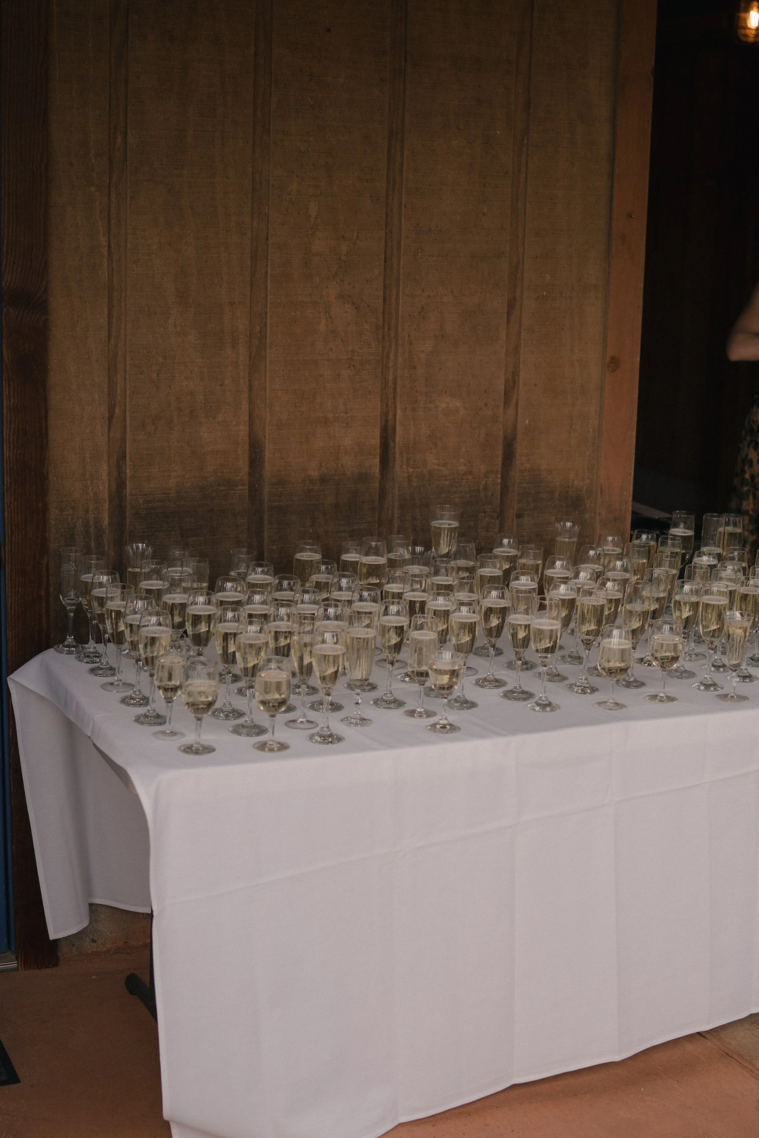 Table covered with a white tablecloth, set with numerous champagne glasses filled with sparkling wine or champagne, against a wooden paneled wall.