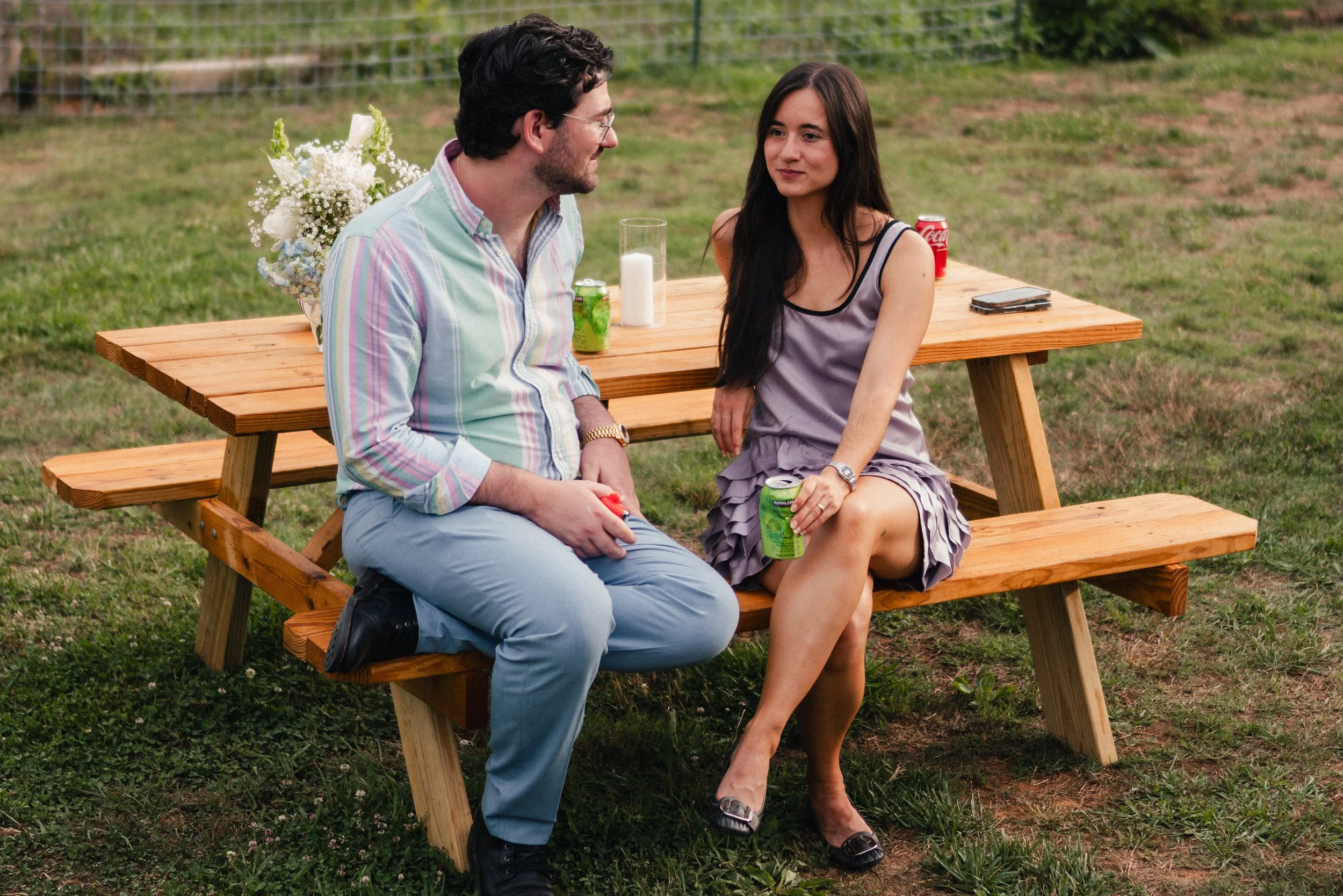 A man and woman sitting at a wooden picnic table outdoors, engaged in conversation with drinks and a smartphone on the table, with greenery in the background.