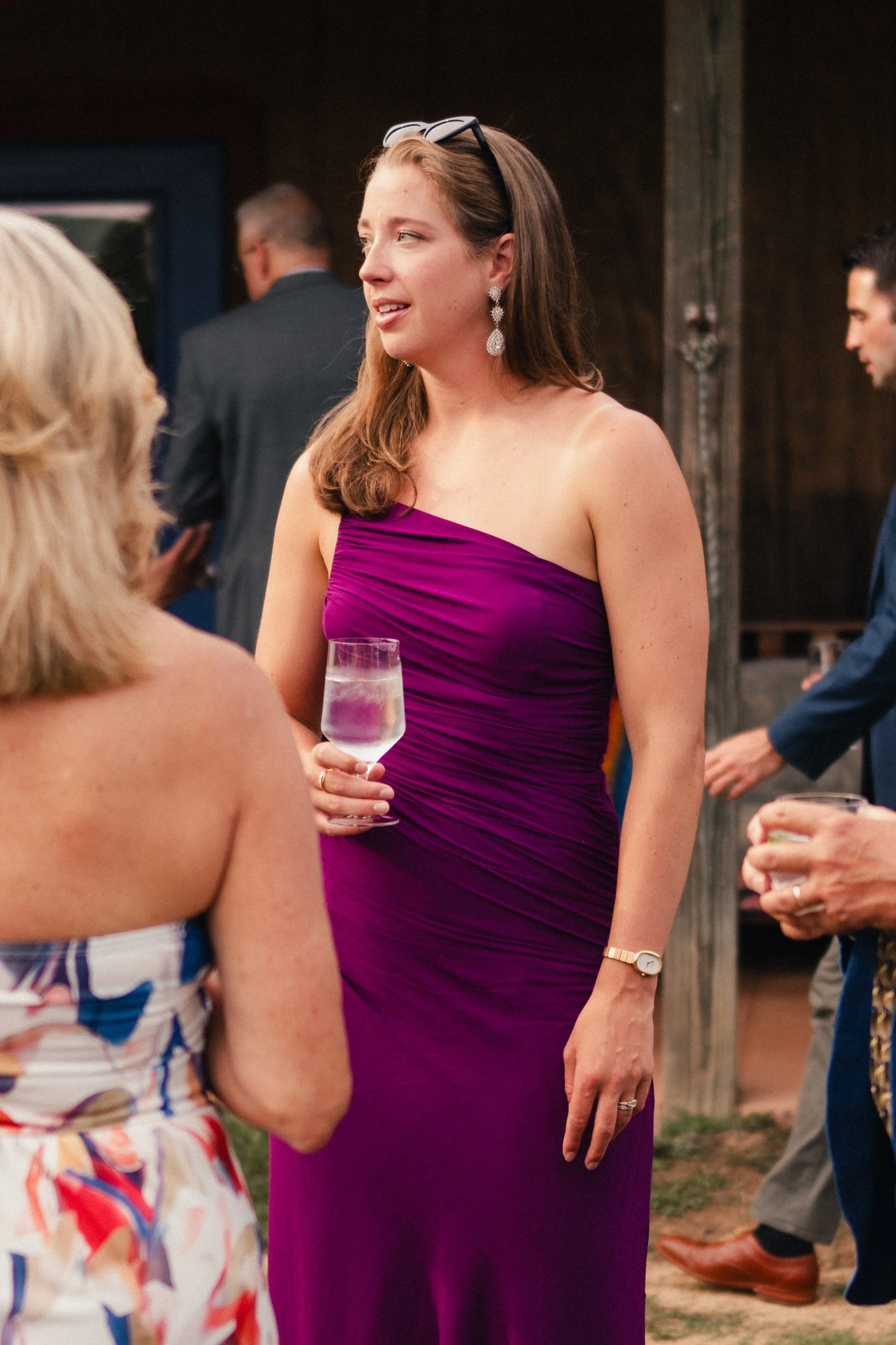 A woman in a purple dress holding a glass of white wine, attending a social event or celebration, with other guests in the background.