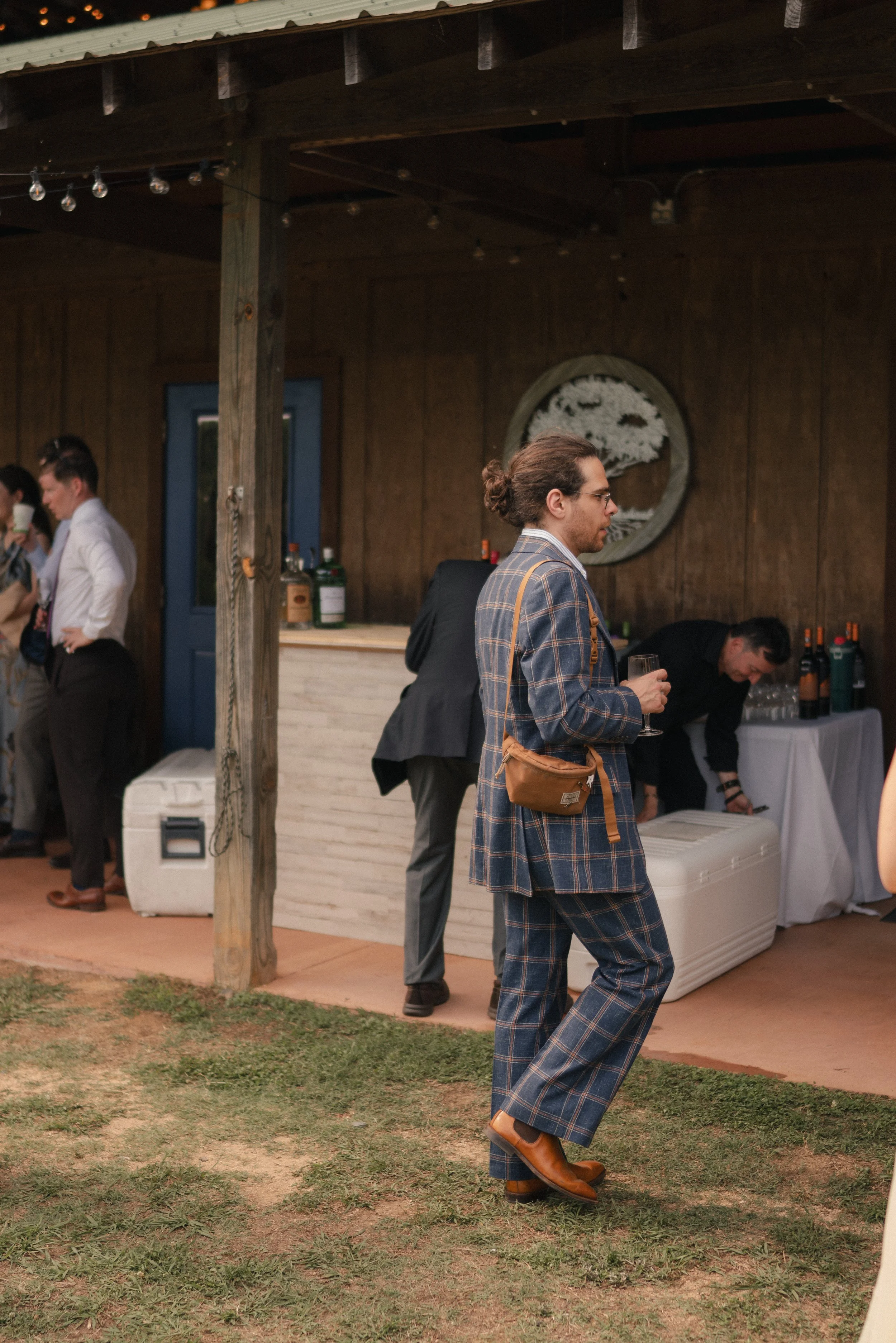 Man in plaid suit and glasses holding a drink, walking outdoors during a social gathering under a wooden roof, with other guests and a bar area in the background.