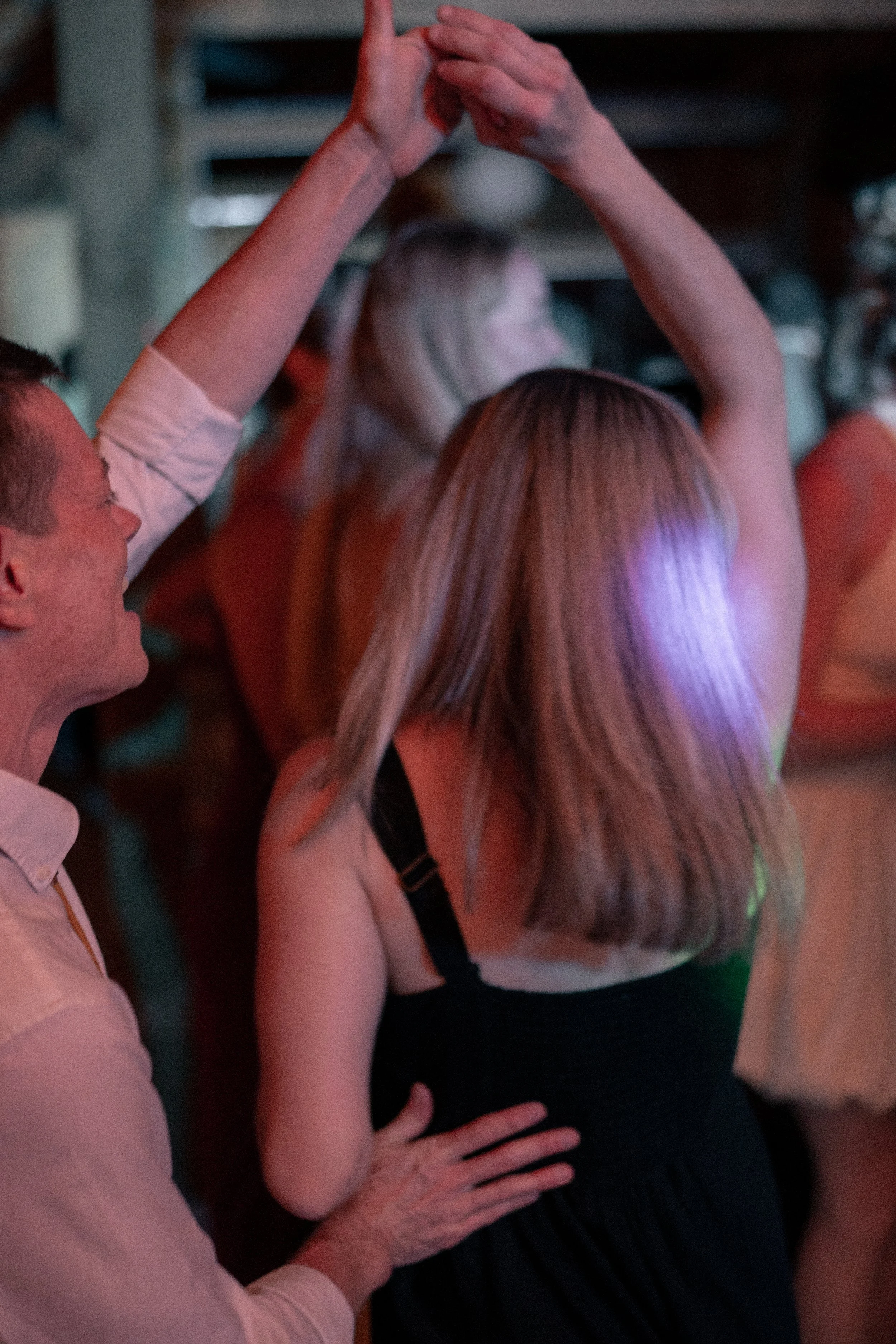 A woman with long blonde hair wearing a black top is dancing at a party or social gathering, visible from behind. A man in a light shirt is next to her, with one of his hands on her waist, and the woman’s hand is holding the man’s hand above their he